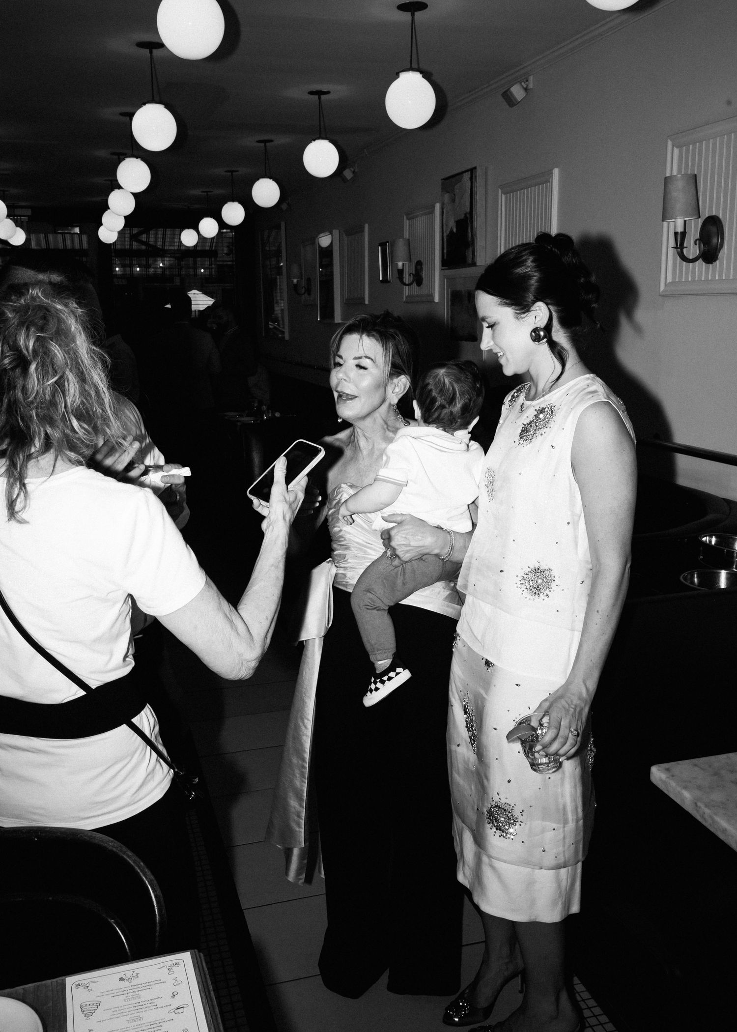 A black and white photo showing people socializing at an event with spherical pendant lights overhead.