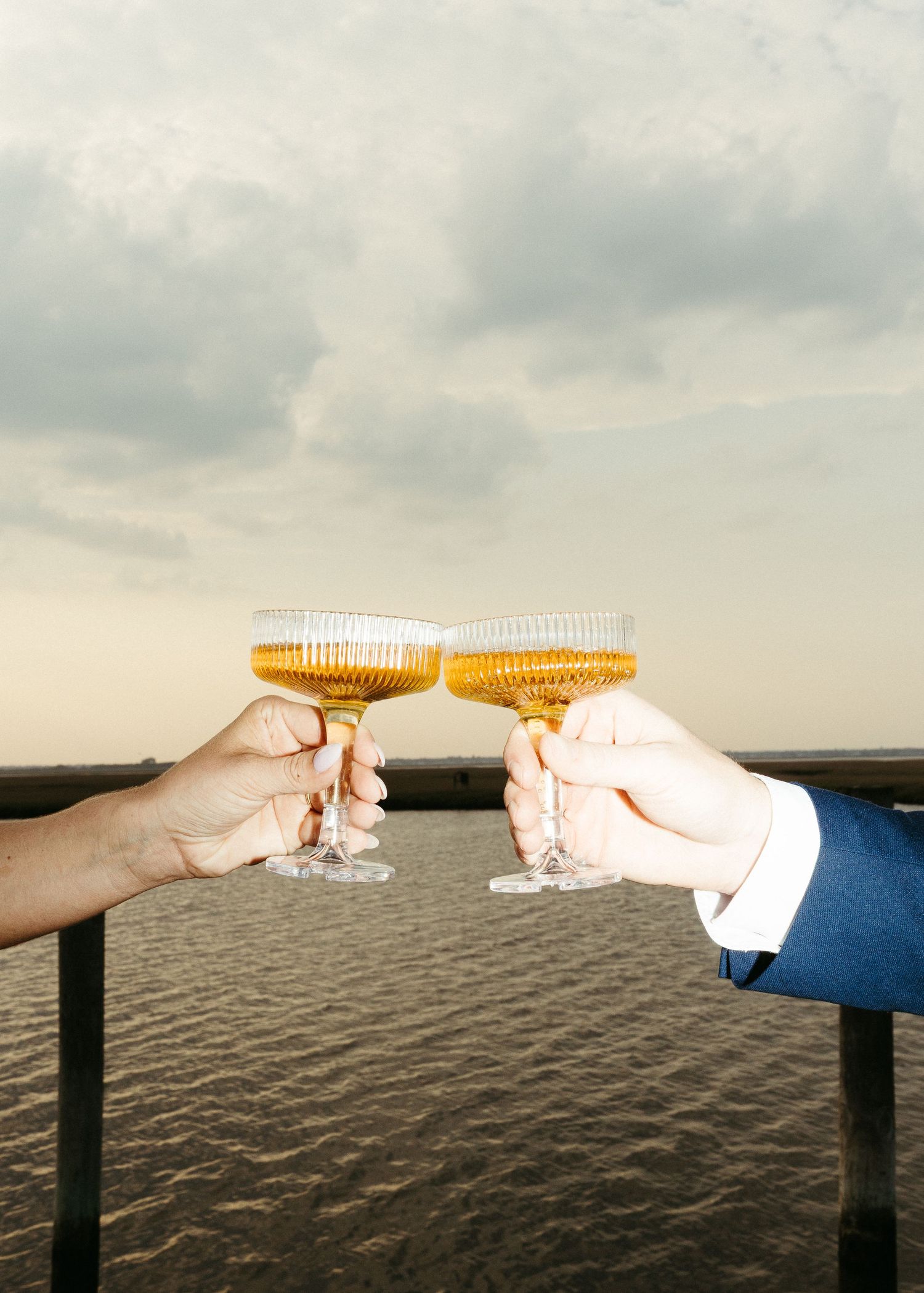 Two hands toasting champagne glasses against a waterfront sunset backdrop.