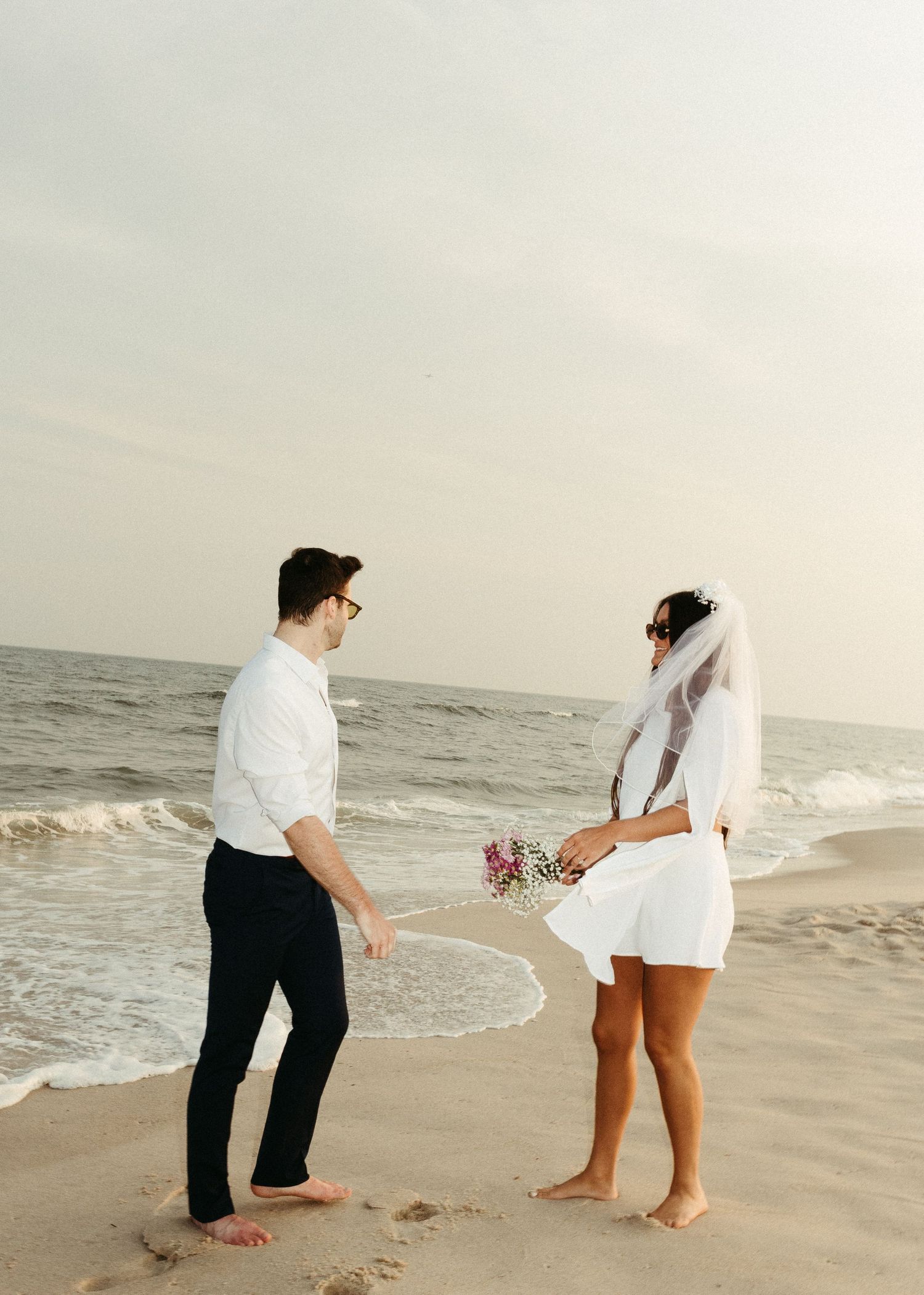 A couple in wedding attire stands together on a beach near the ocean waves.