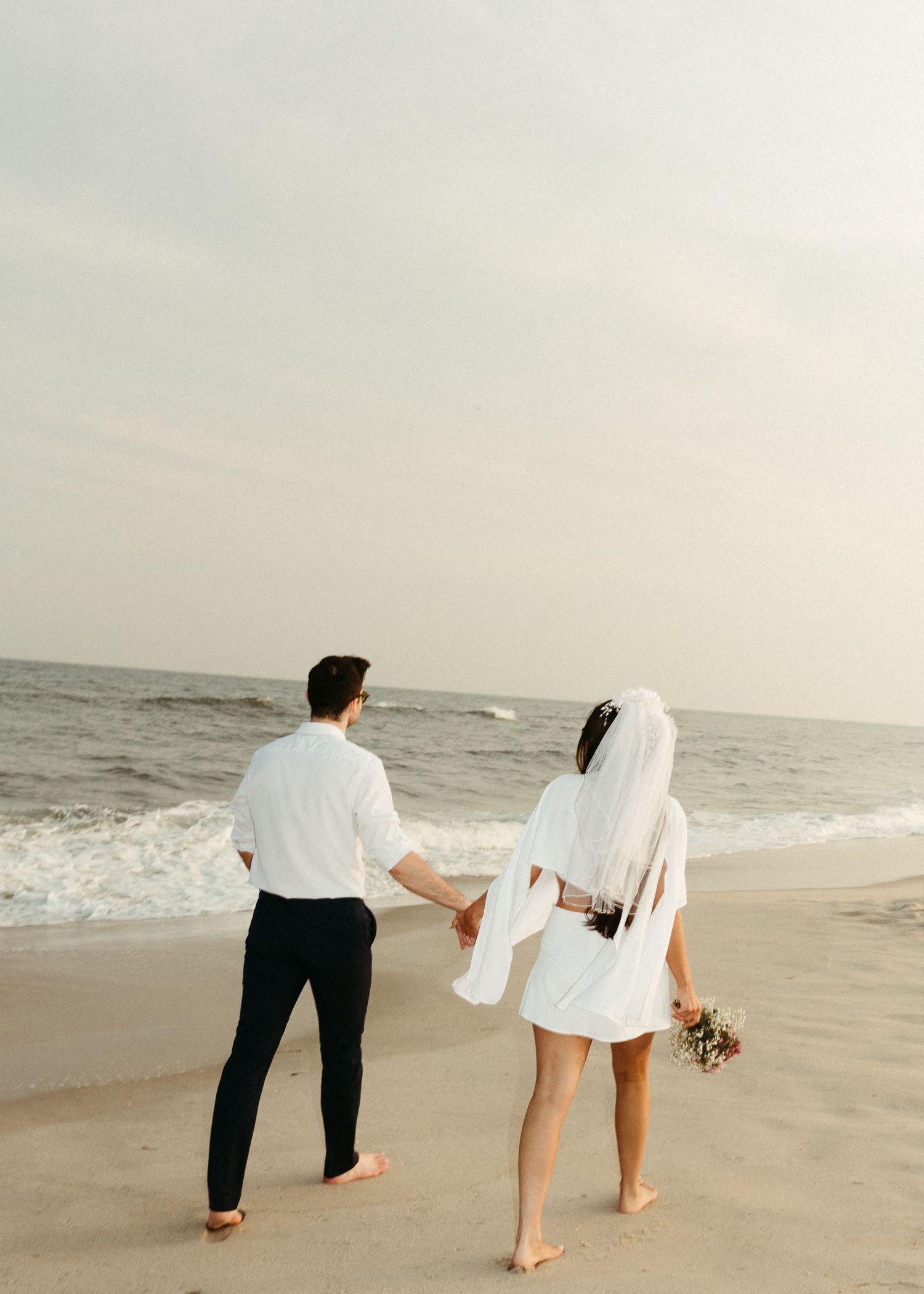 A romantic beach wedding couple walks barefoot along the shoreline holding hands.