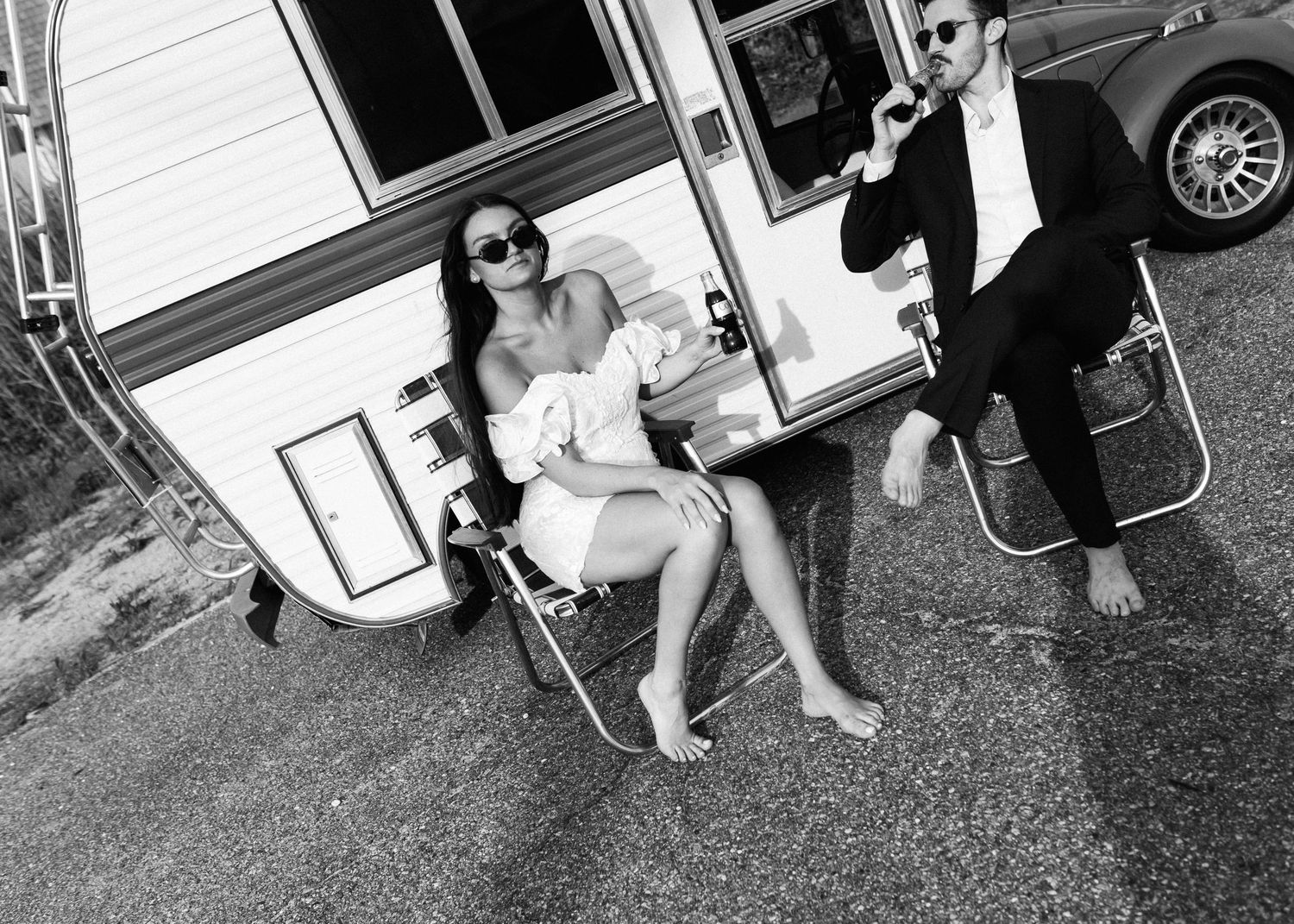 Black and white photo of a couple enjoying drinks outside their vintage camper trailer.