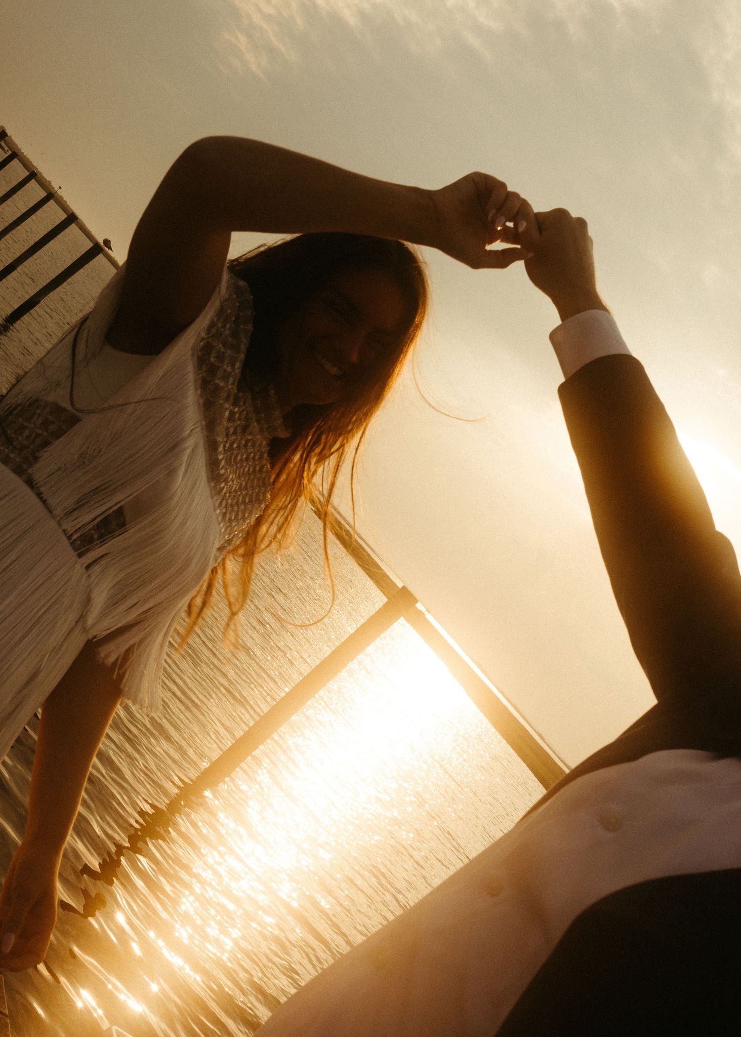 A couple dances on a pier with sunlit water sparkling behind them.