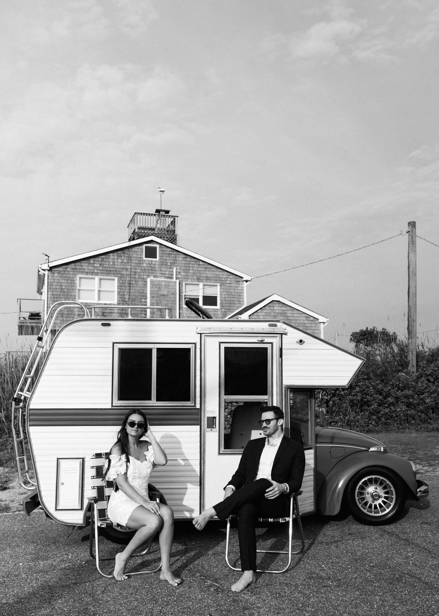 Black and white photo of couple relaxing in chairs beside retro camper and classic car.