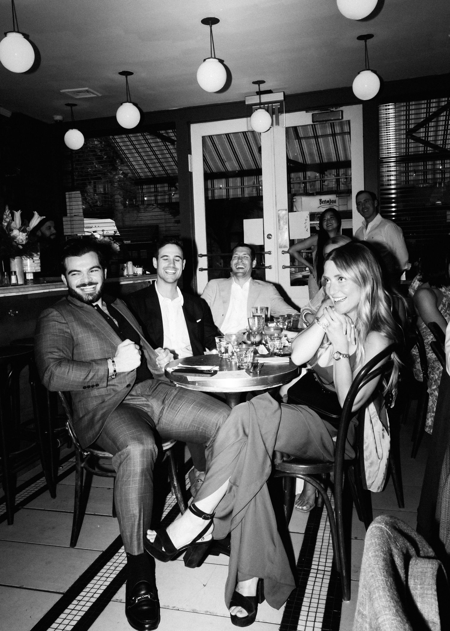Black and white restaurant scene with guests seated at table under pendant lighting.