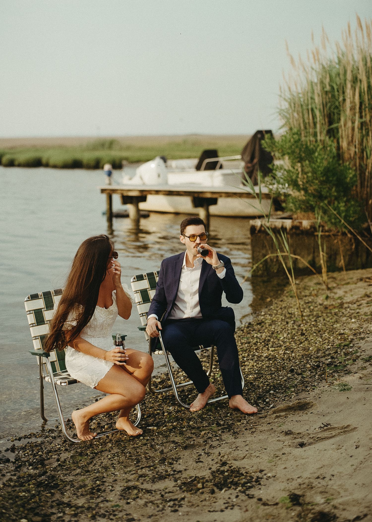 People relaxing on a rocky beach shoreline with tall reeds and a wooden dock in the background.