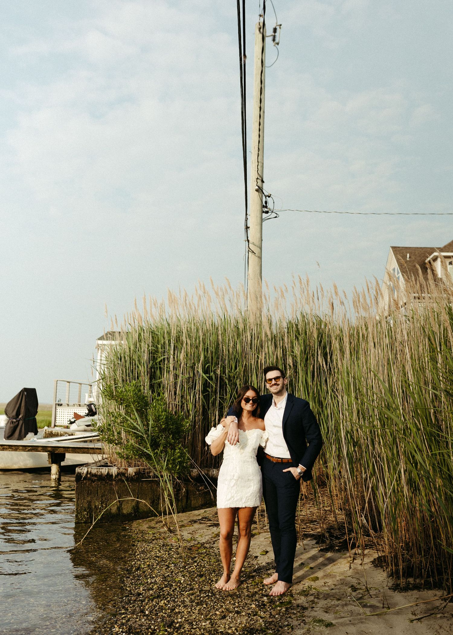 A couple poses by marsh grasses near a boat dock on a sunny day.