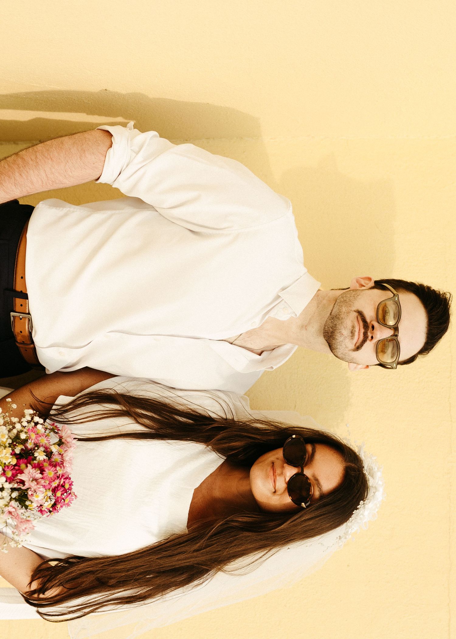 A couple in matching white outfits poses from above against a yellow background.