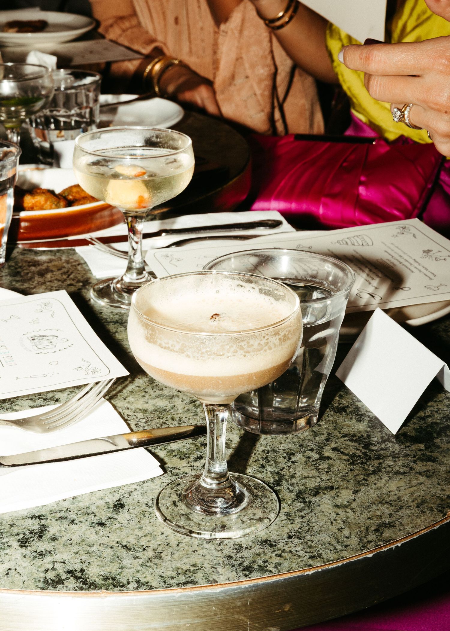 Frothy cocktail in coupe glass on marble table with menus and dining items.