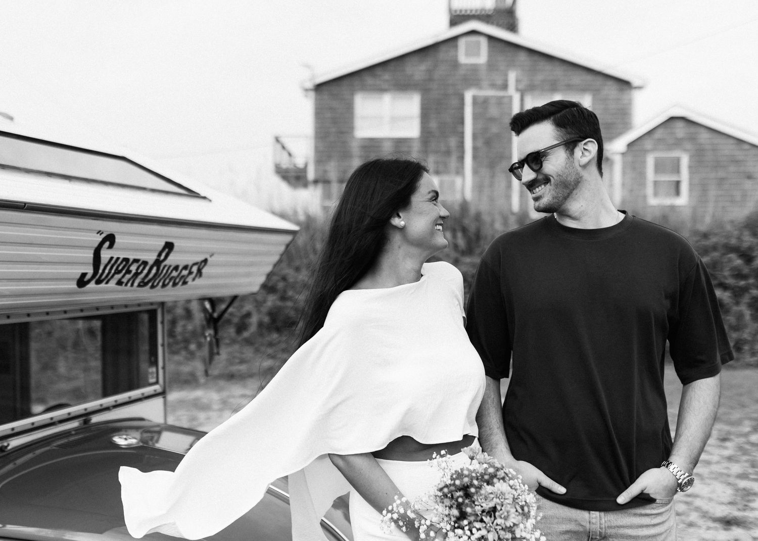 A couple poses romantically by a vintage trailer in a black and white photo holding flowers.