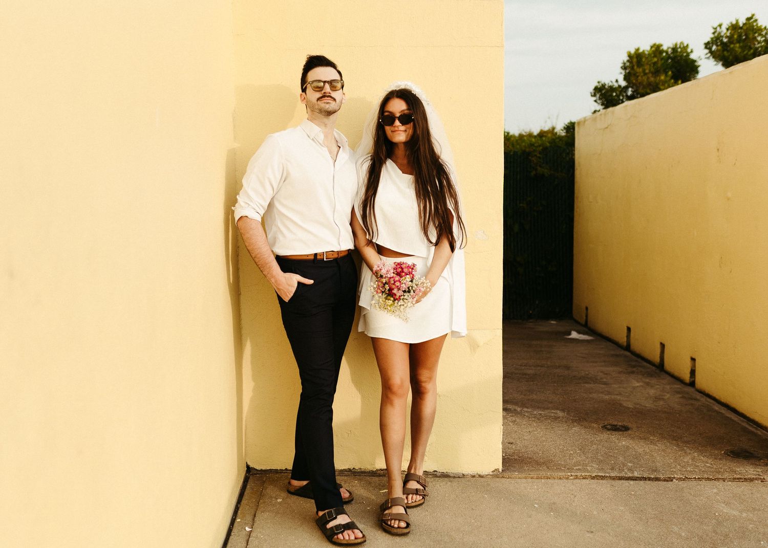 Stylish couple poses against yellow wall, wearing white and matching casual outfits with sandals outdoors.