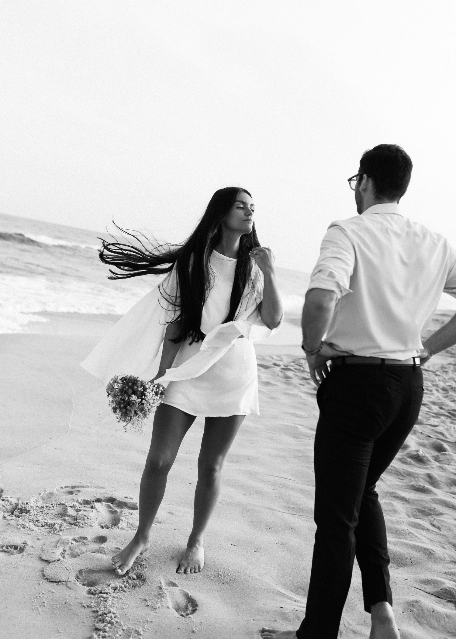 A candid moment of a couple in white outfits enjoying a breezy day at the beach.