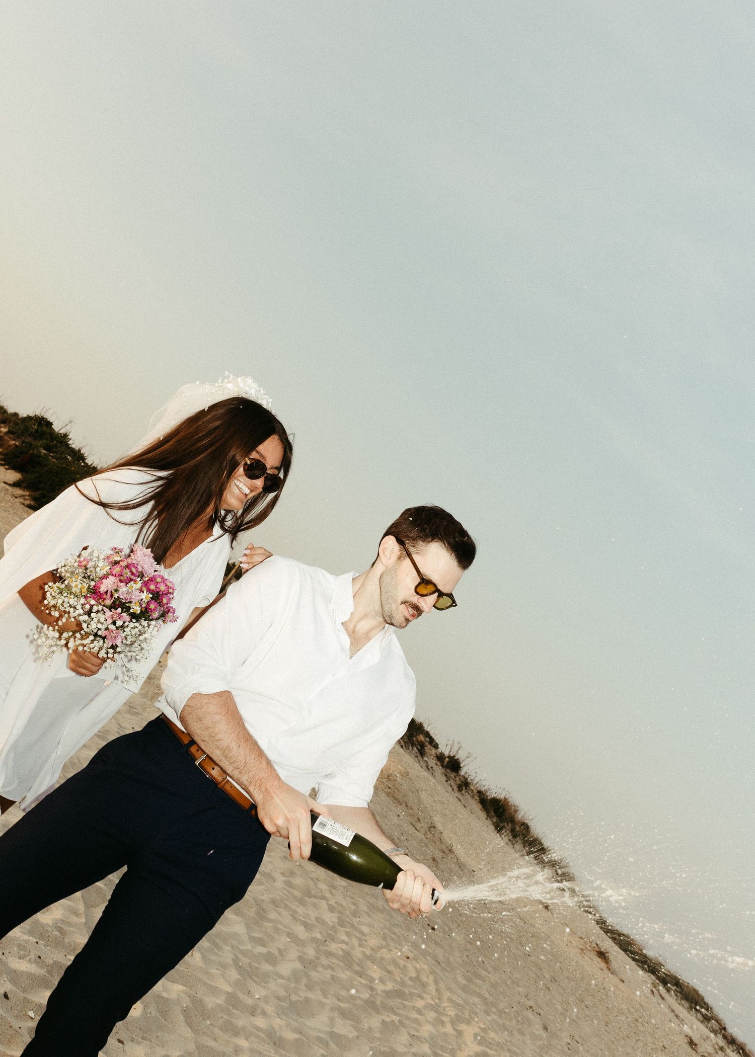 A couple in matching white outfits opens champagne during their beach photoshoot.