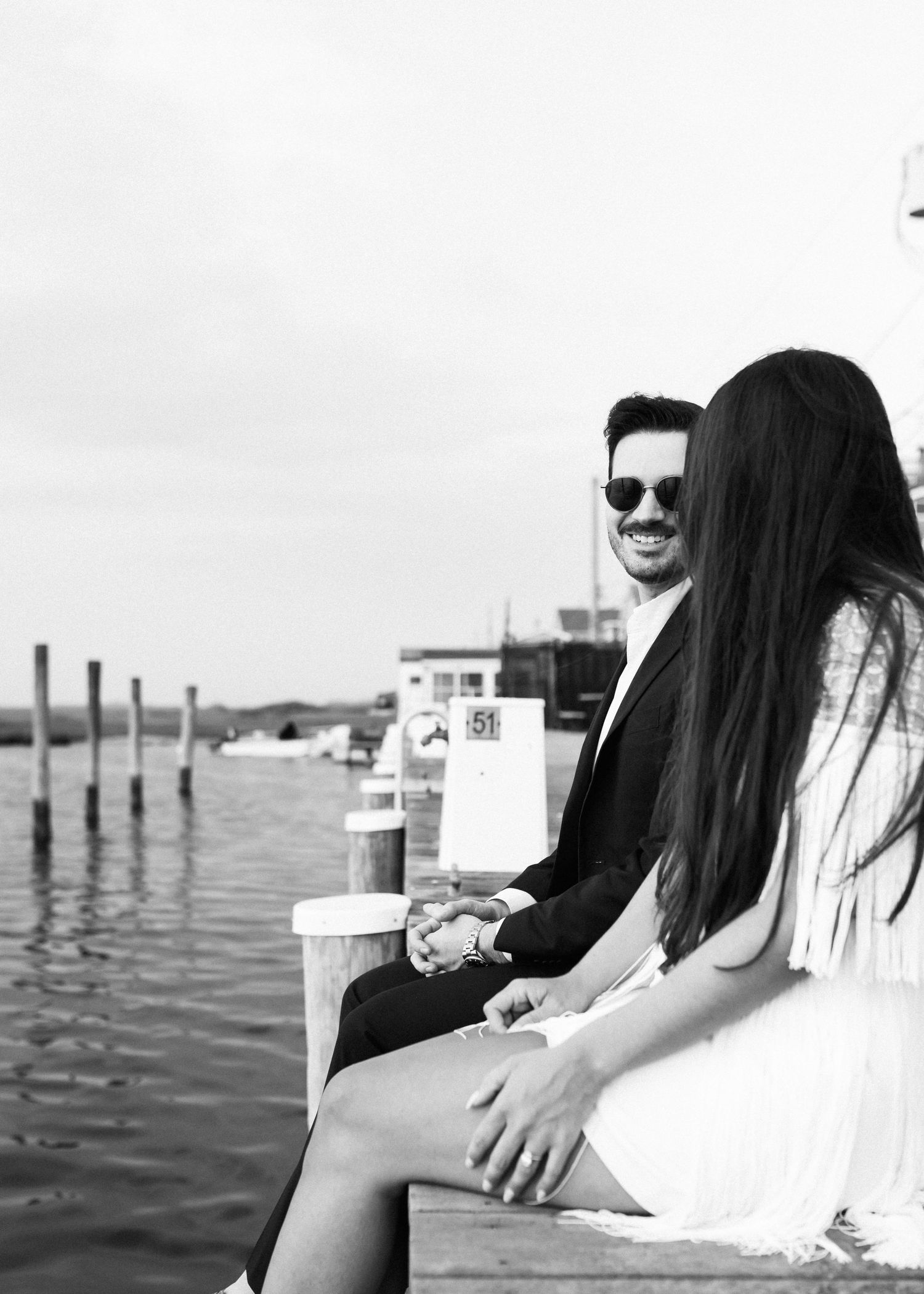A couple sits together on a dock overlooking a marina with wooden posts and boats in the background.