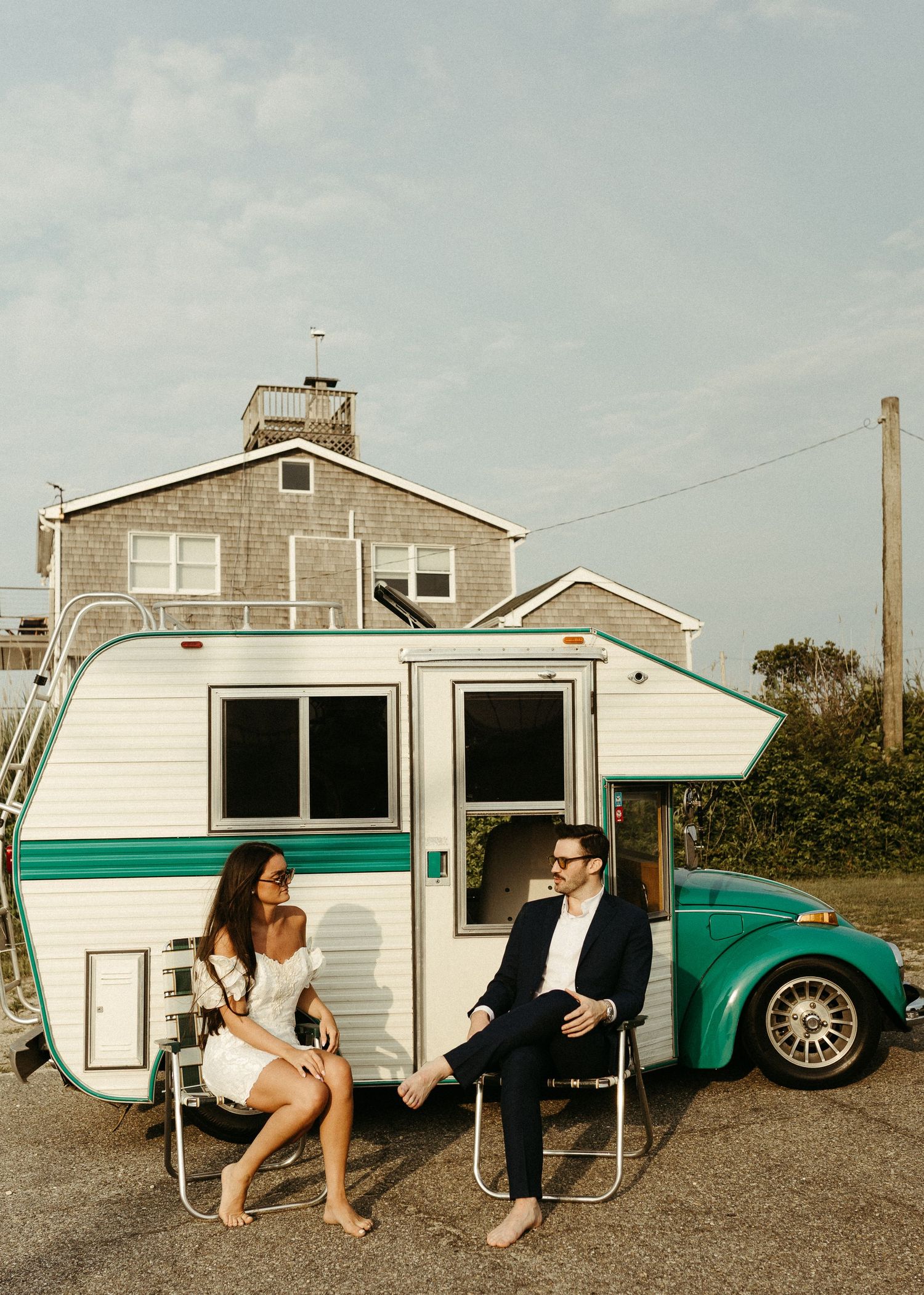 Couple relaxing in folding chairs next to a vintage teal and white camper trailer with a house in the background.