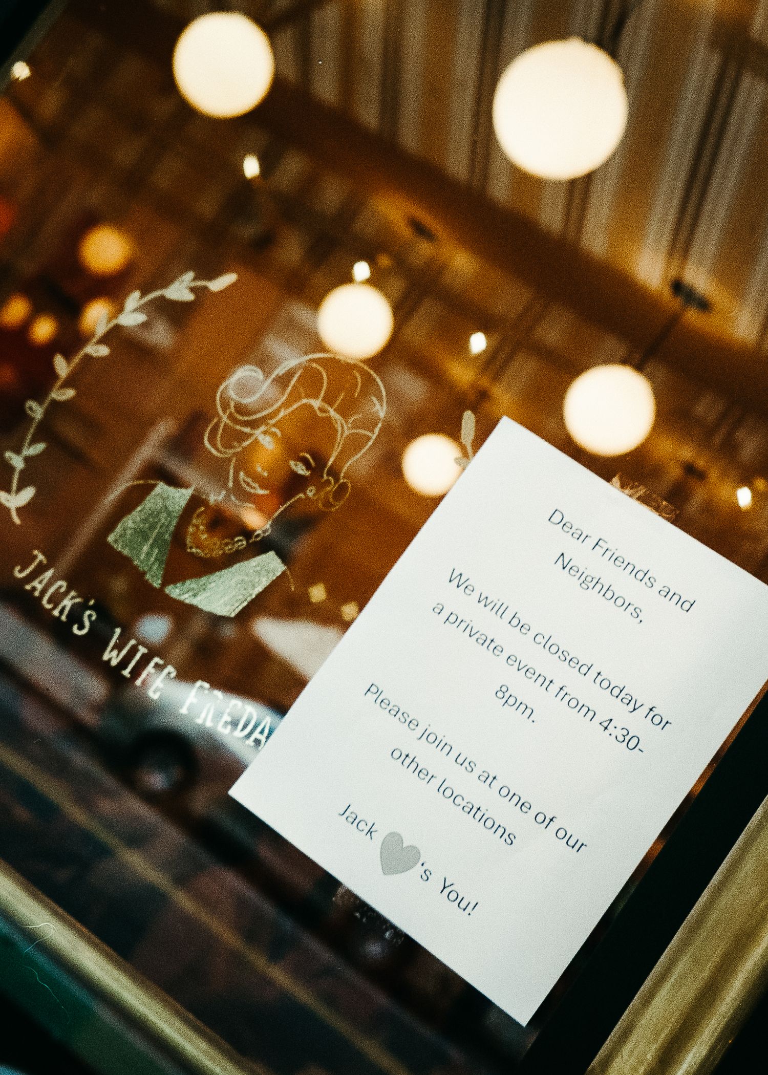 Window sign with handwritten note and glowing pendant lights reflected in glass.
