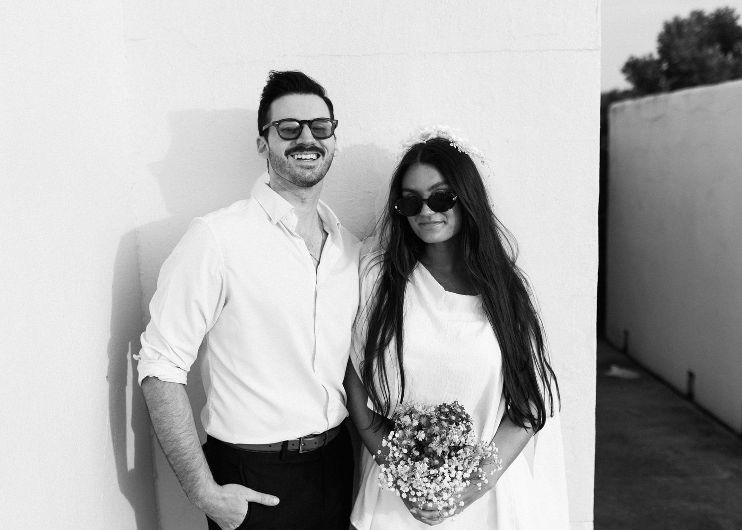 Black and white photo of couple in white outfits against wall, holding floral bouquet.