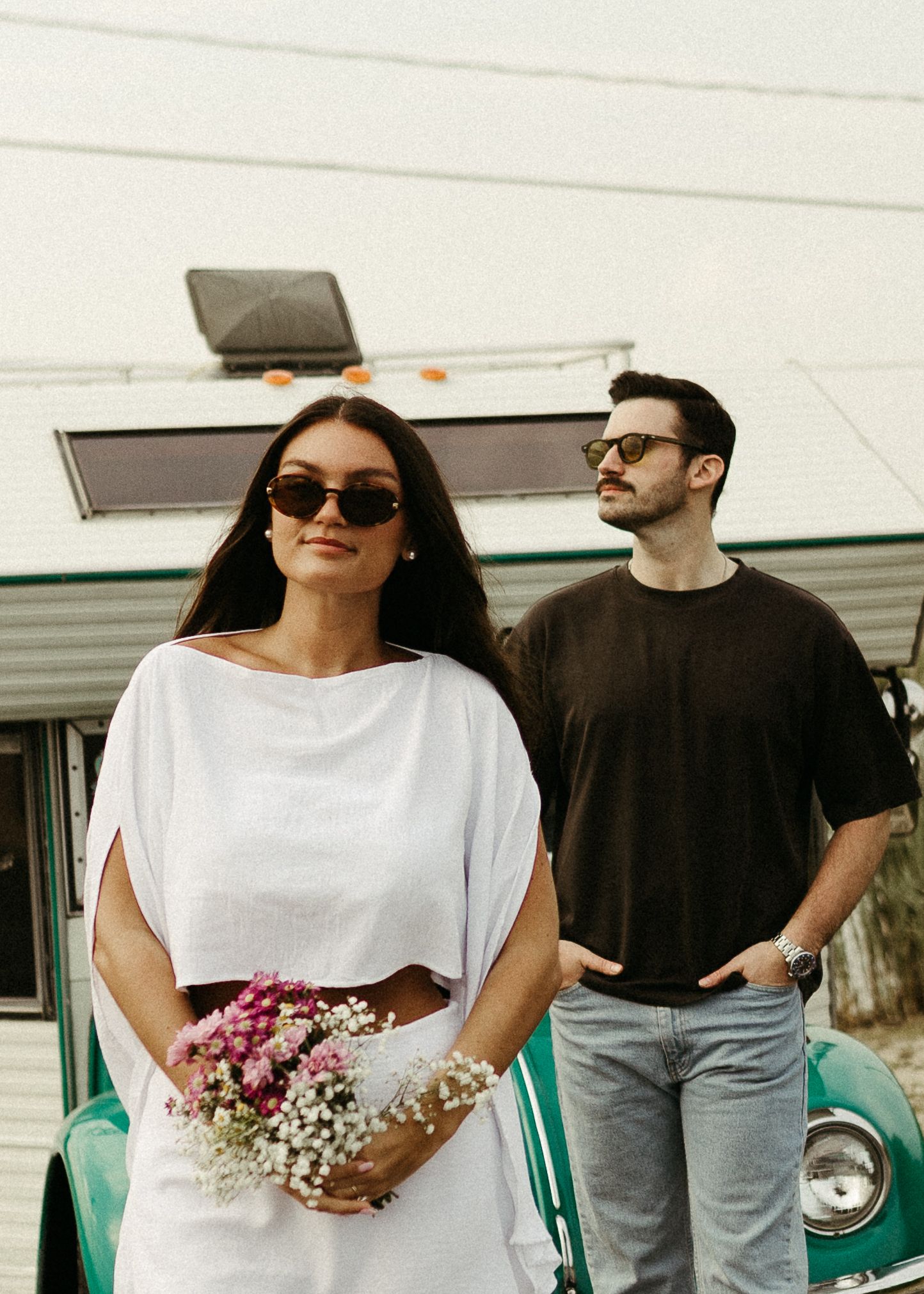 A couple stands by a retro camper with flowers and sunglasses in casual summer attire.