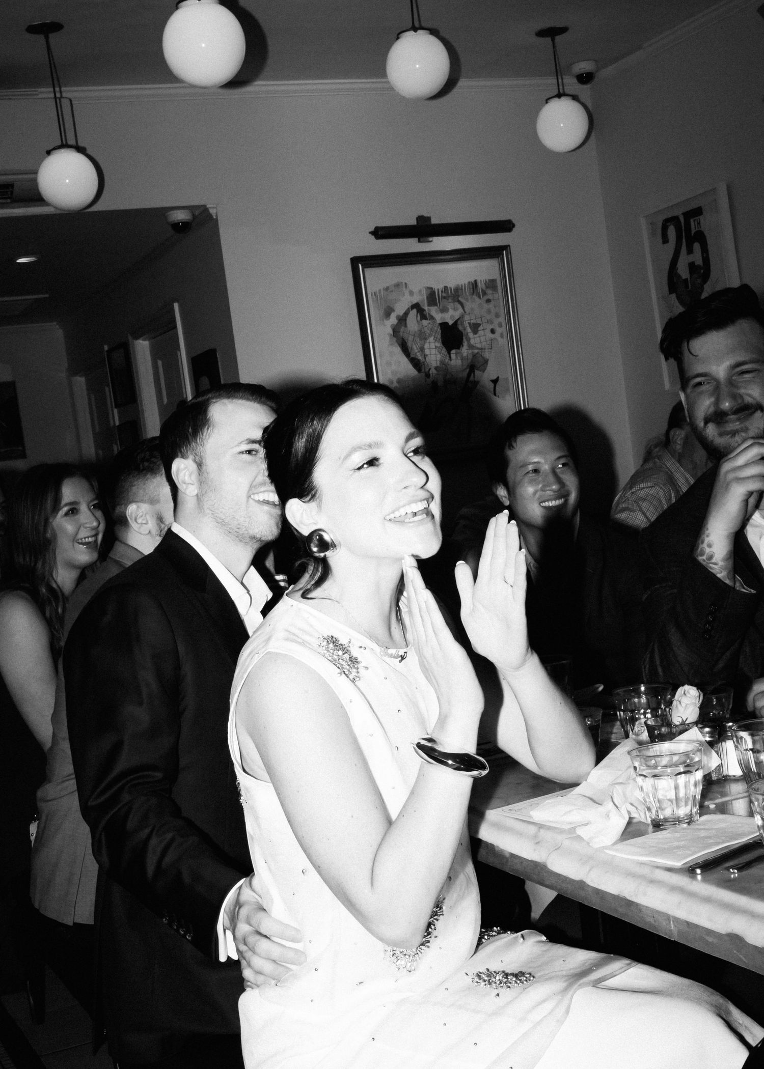Black and white photo of dinner party guests enjoying themselves under round ceiling lights.