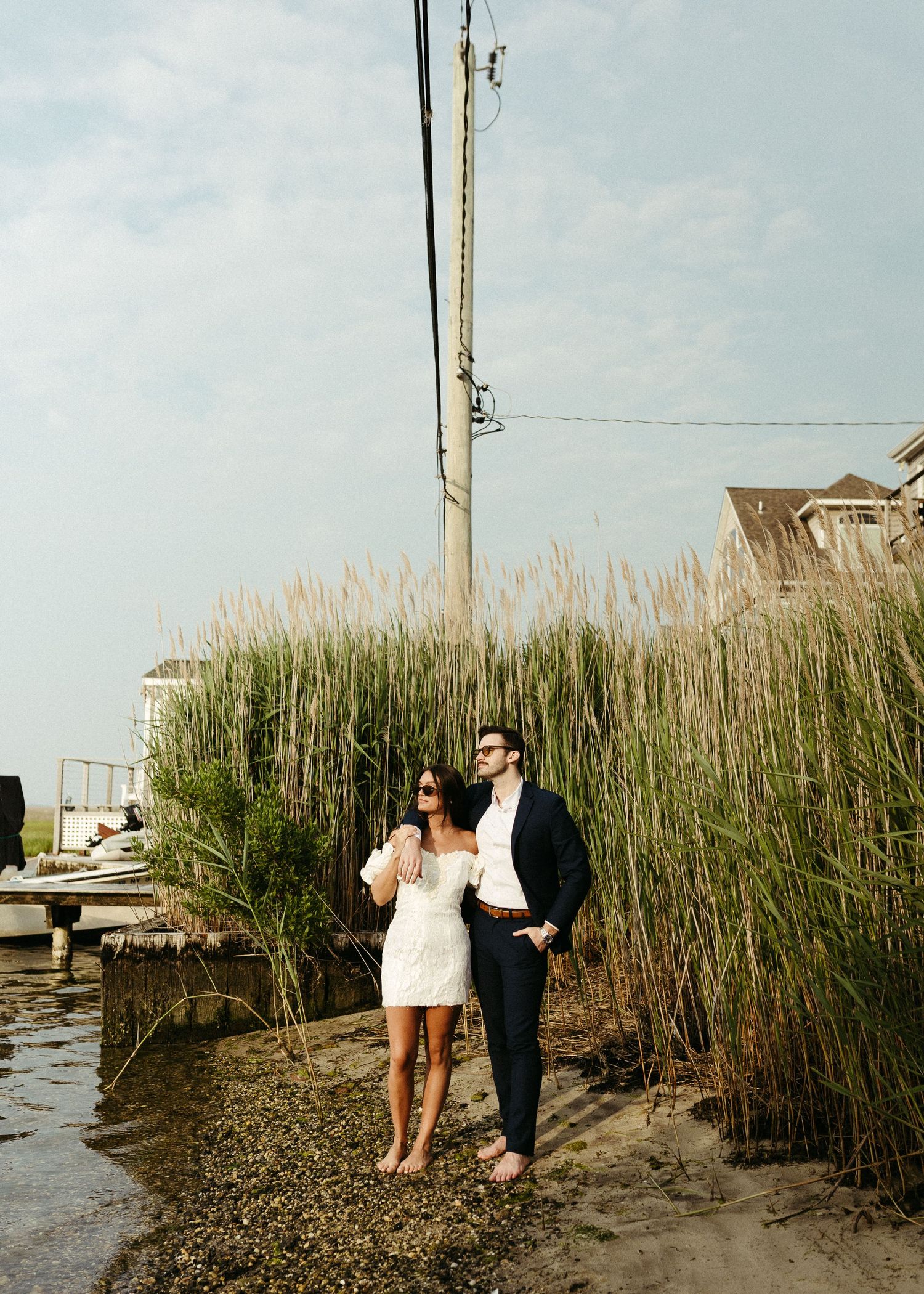 A couple stands near tall reeds by the waterfront during a coastal photoshoot.