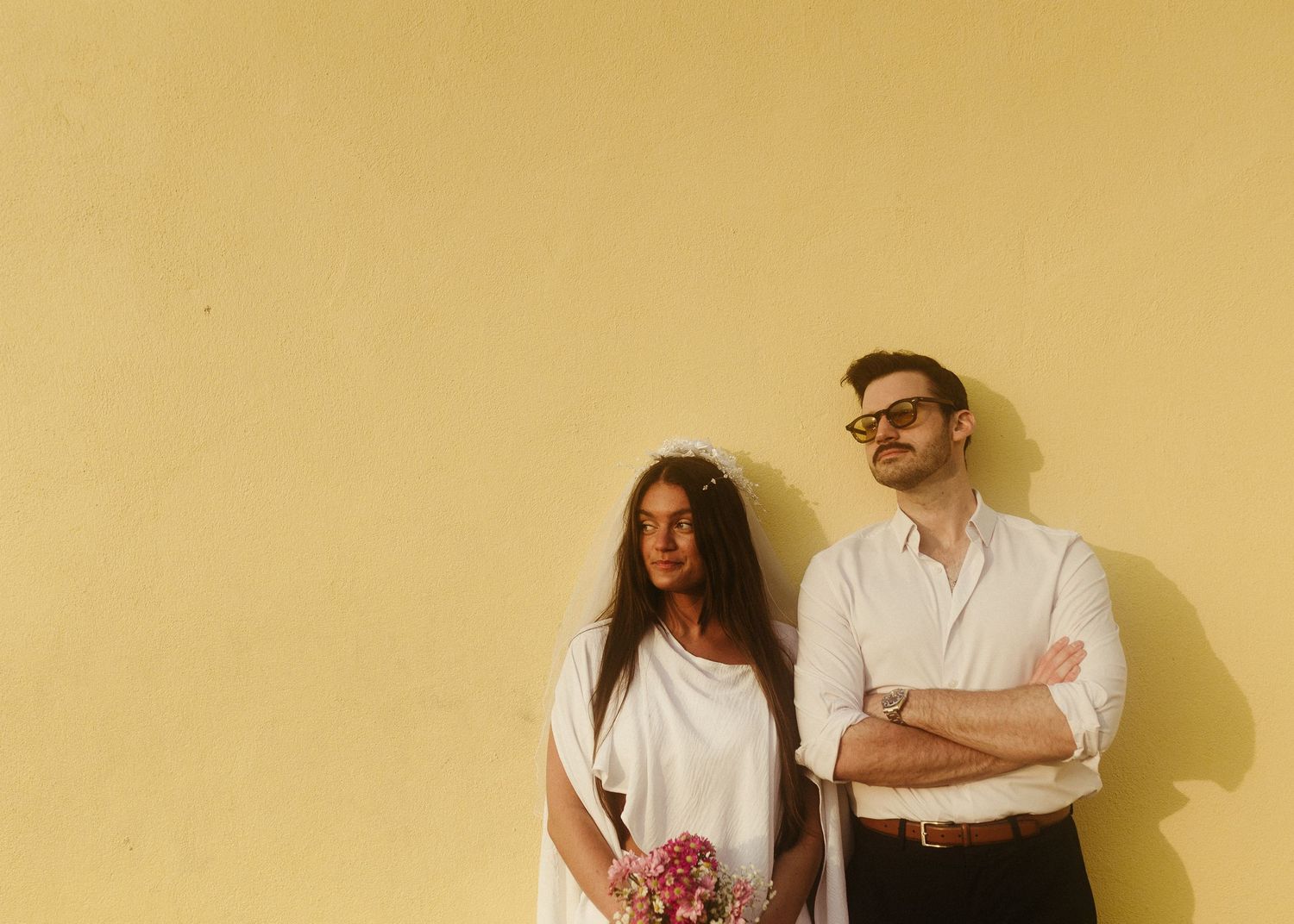 Minimal portrait of couple against yellow wall with bouquet and casual attire.