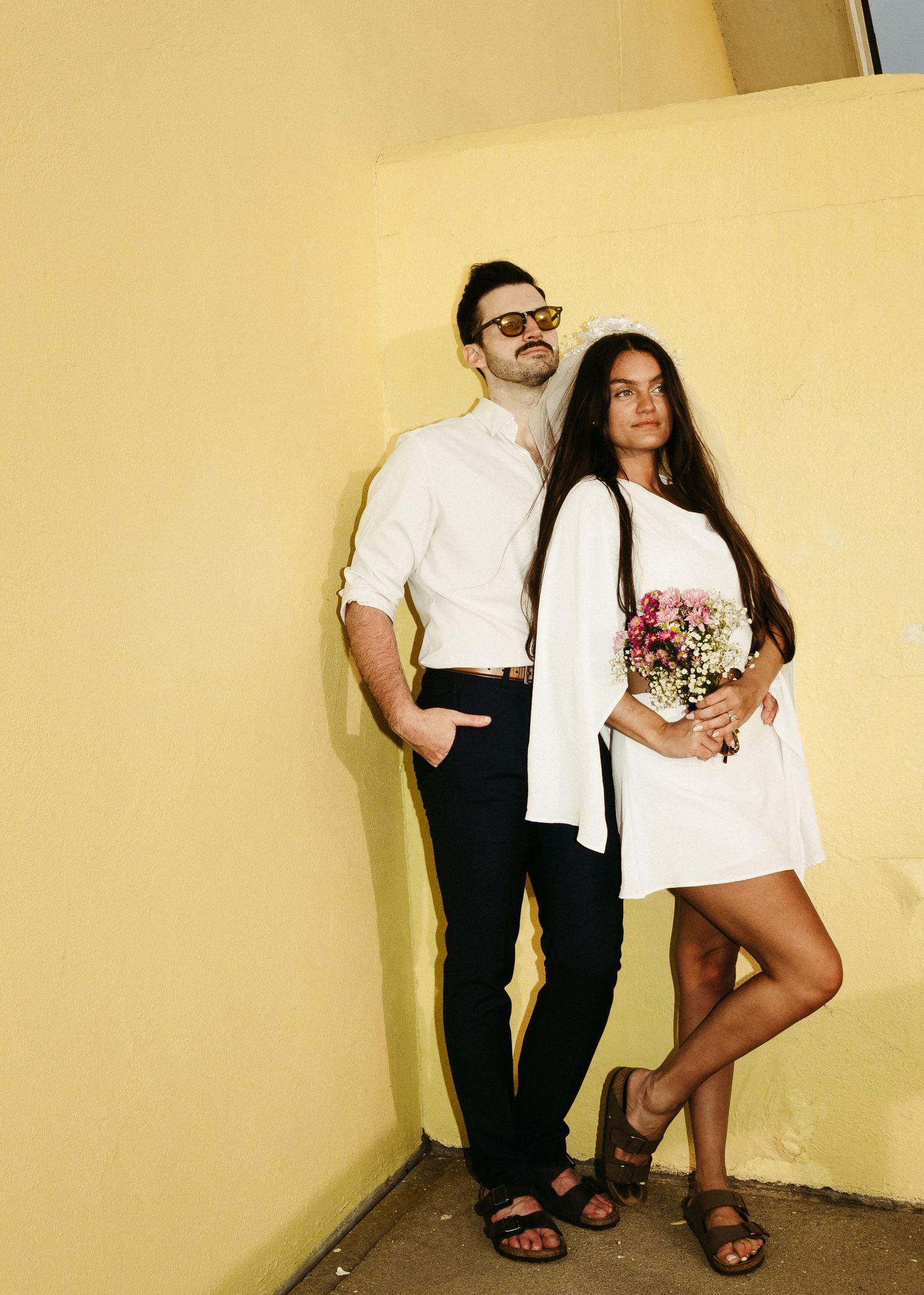 Couple in coordinated white and black outfits pose casually against yellow wall.