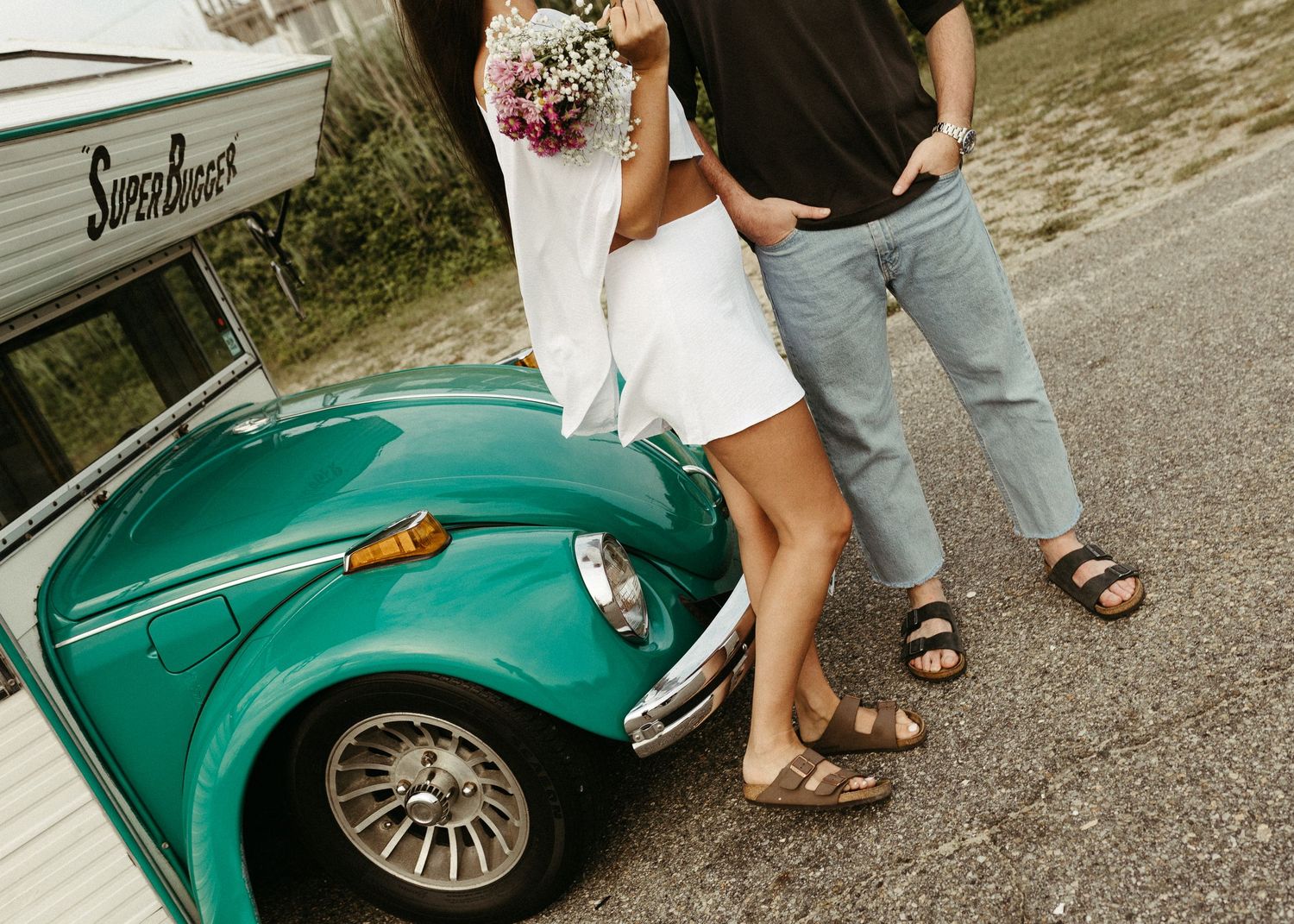 Couple posing with vintage teal Volkswagen Beetle on coastal road.
