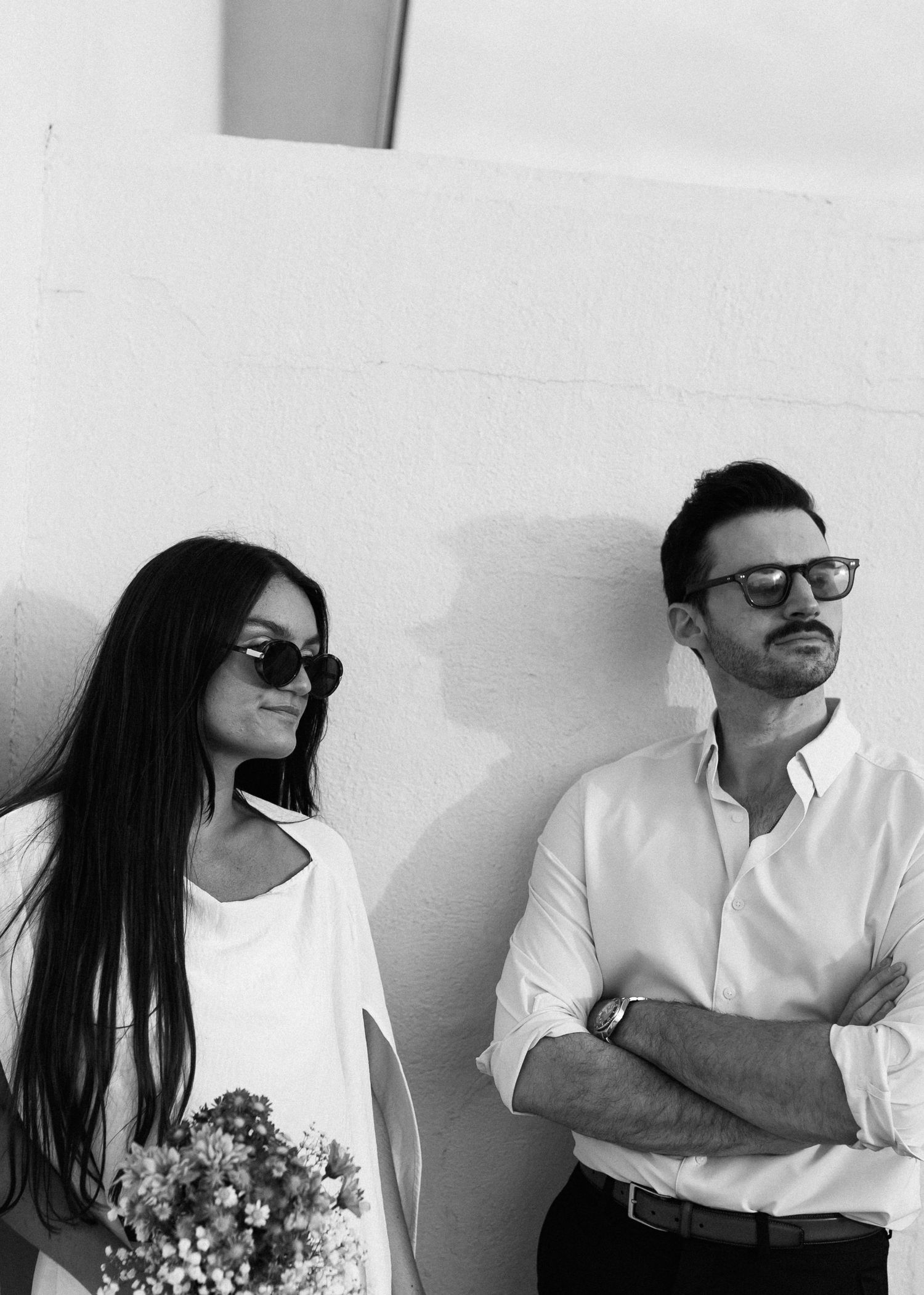 A stylish couple in sunglasses poses against a white wall with a bouquet of pink flowers.