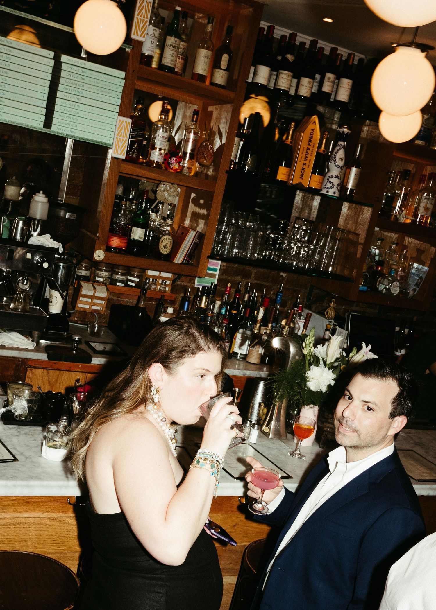 A couple sharing drinks at a well-stocked bar with ambient lighting and glassware.