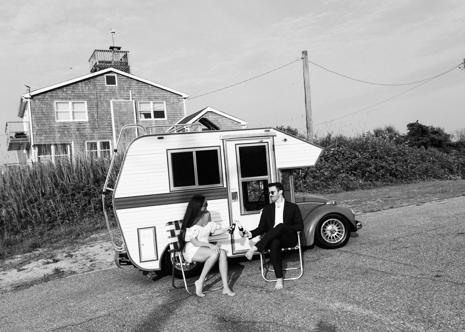 Vintage black and white photo of a retro camper trailer parked near a coastal house.
