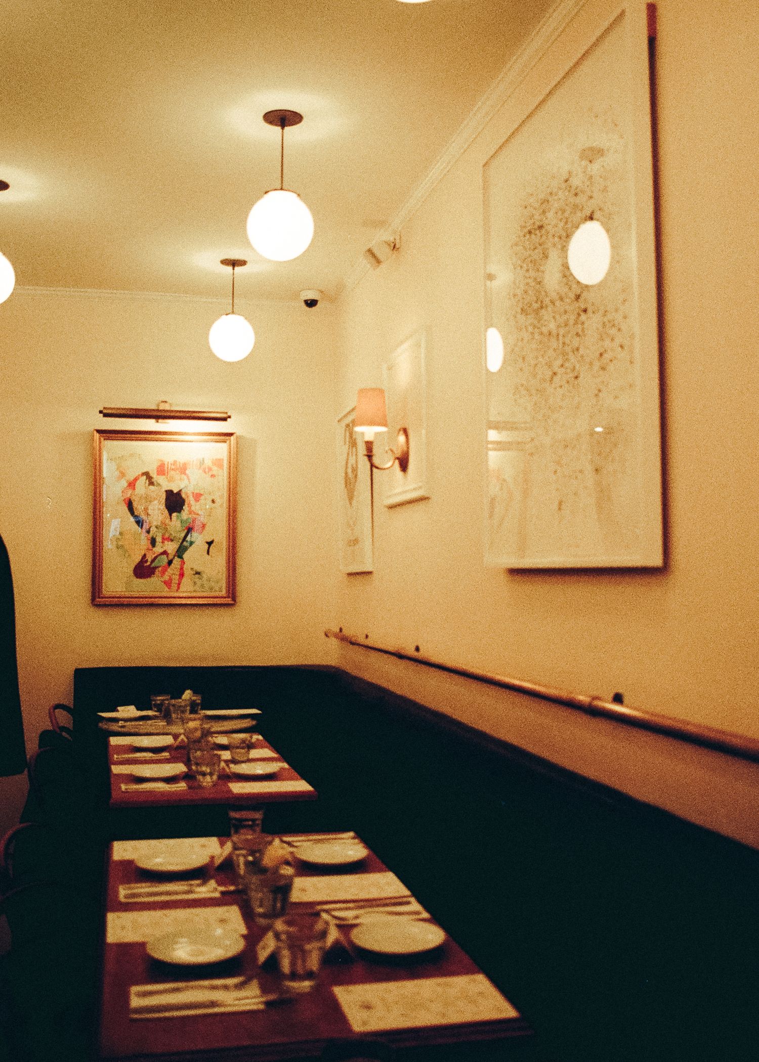 Restaurant interior with globe pendant lights and place settings along a banquette with artwork on cream walls.