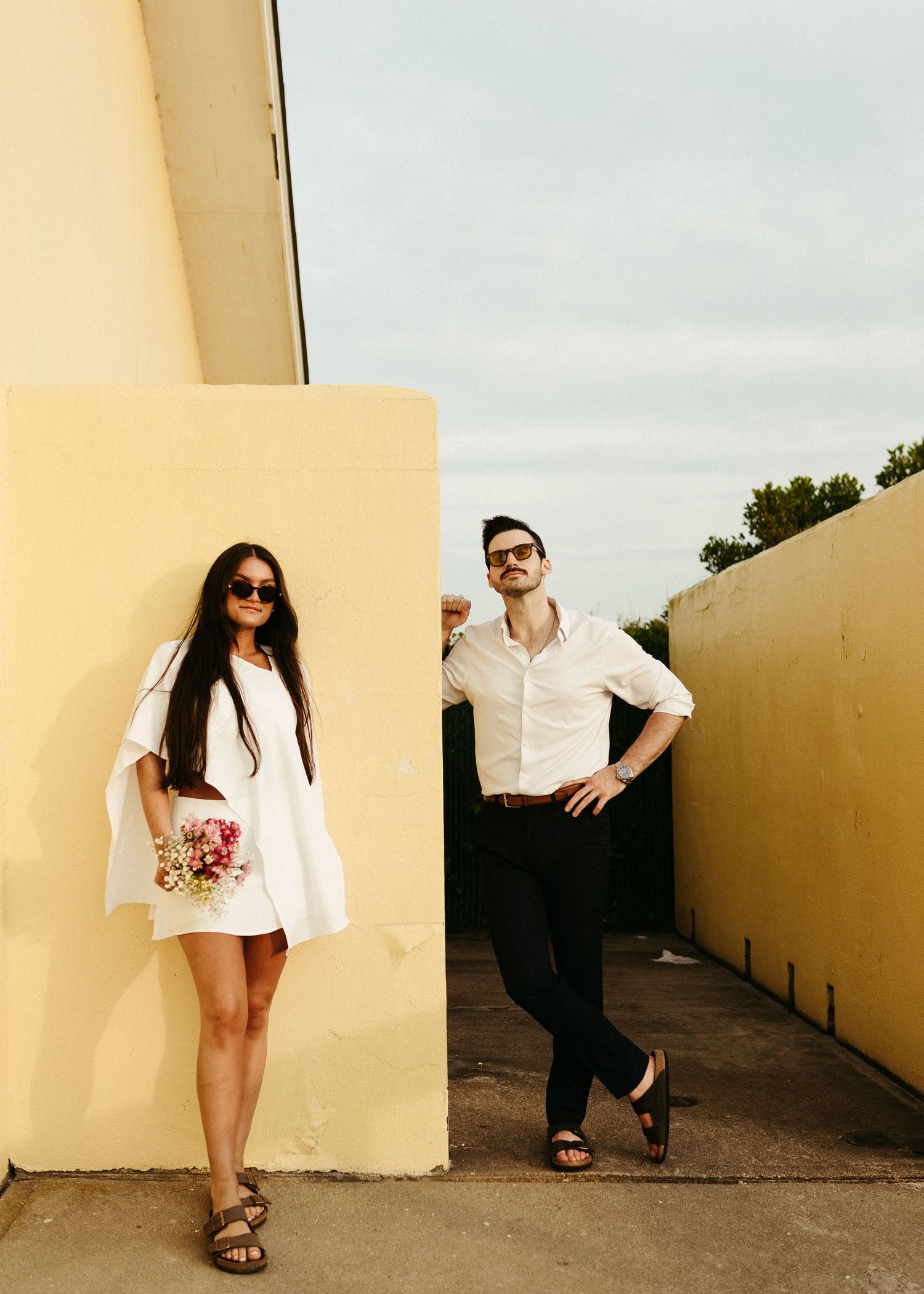 Stylish couple in white outfits posing separately against yellow walls outdoors.