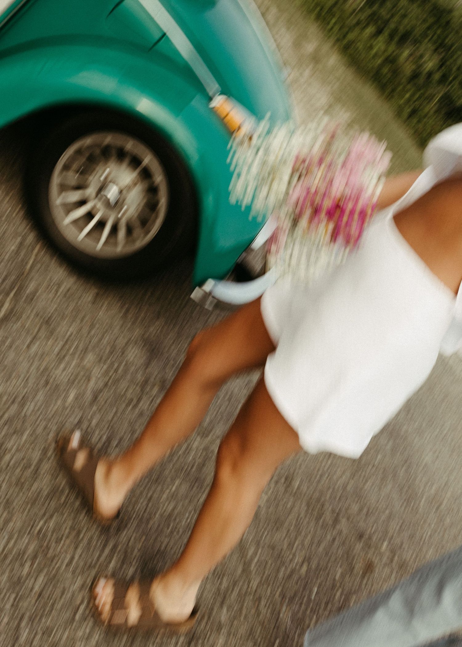 A blurred motion shot of someone in white holding flowers next to a turquoise vintage car.