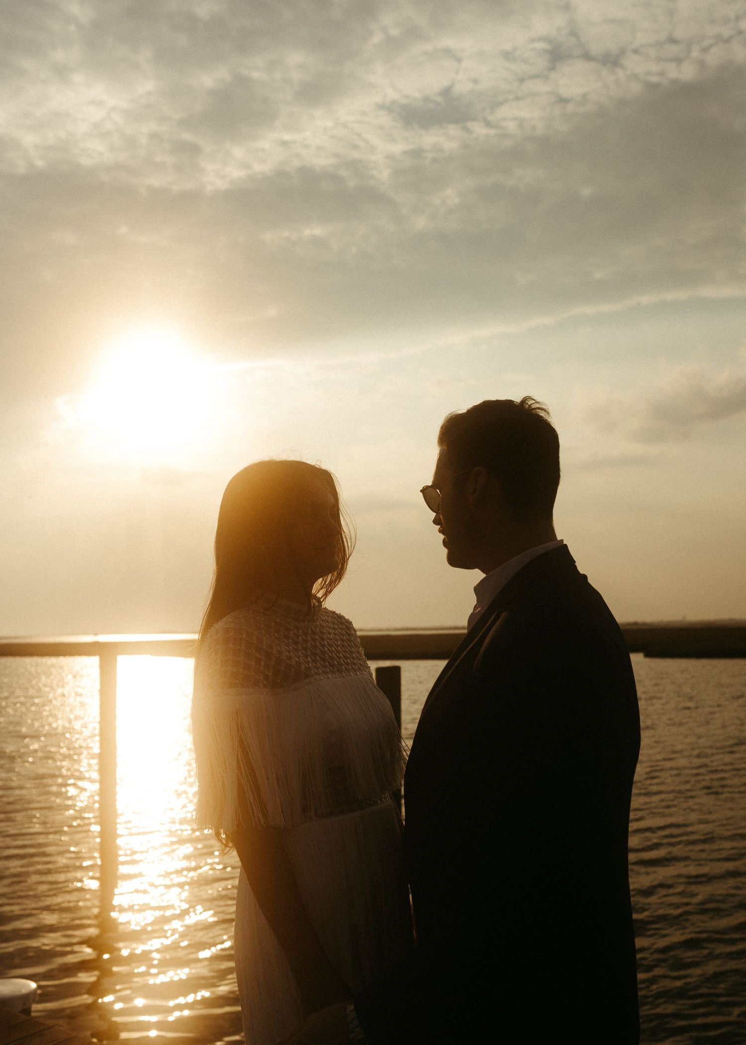 A romantic couple stands in silhouette against a sunset sky over water.