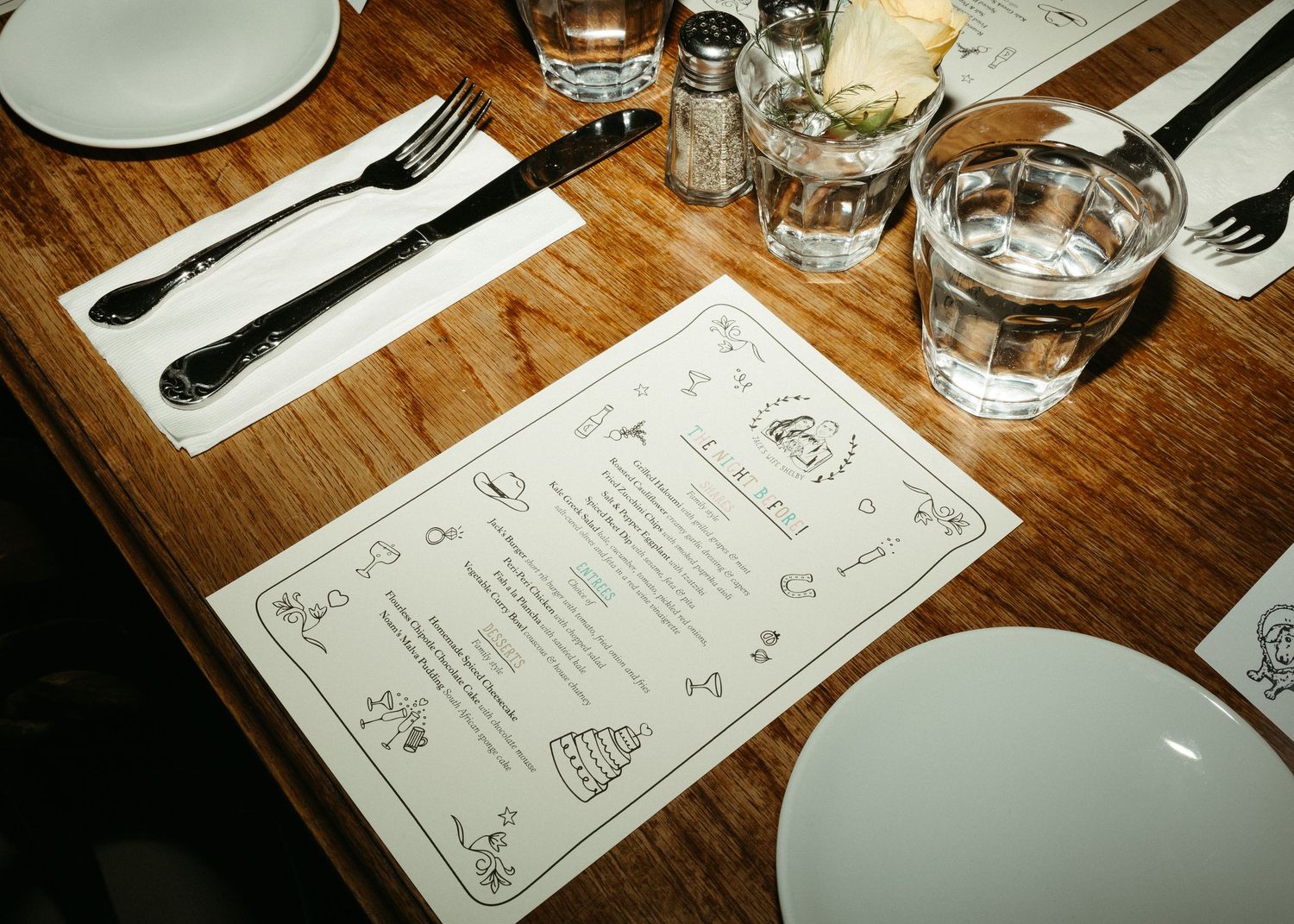 A restaurant table setting with menu cards, white plates, and drinking glasses on a wooden surface.
