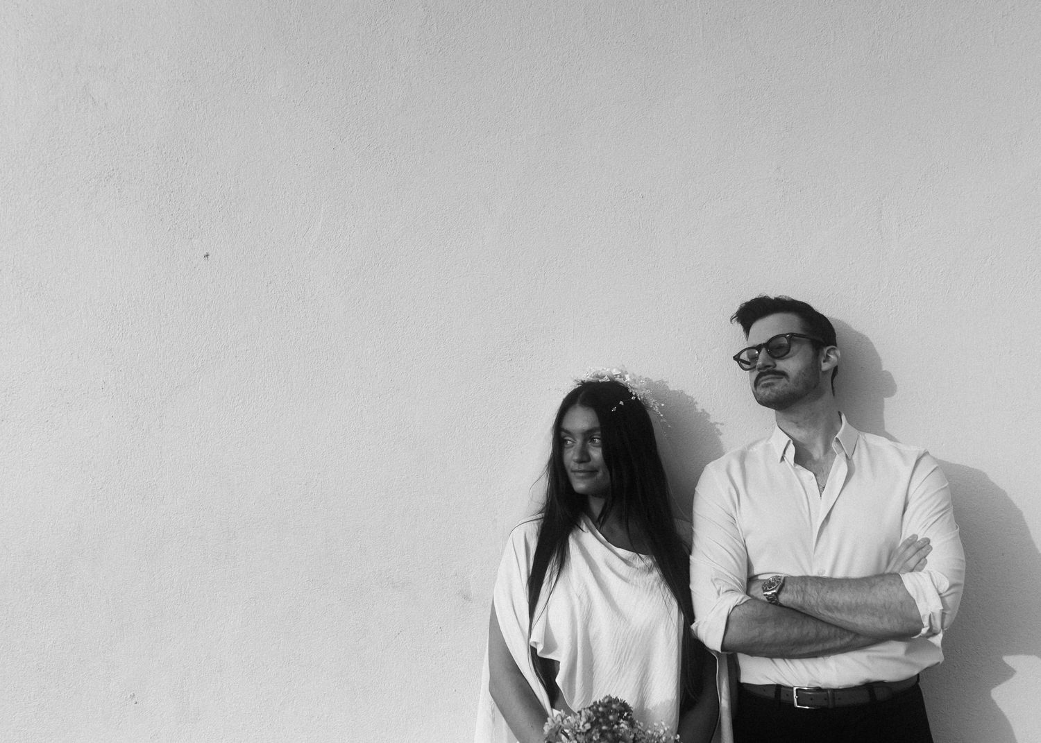 A modern wedding couple poses against a yellow wall in sophisticated attire.
