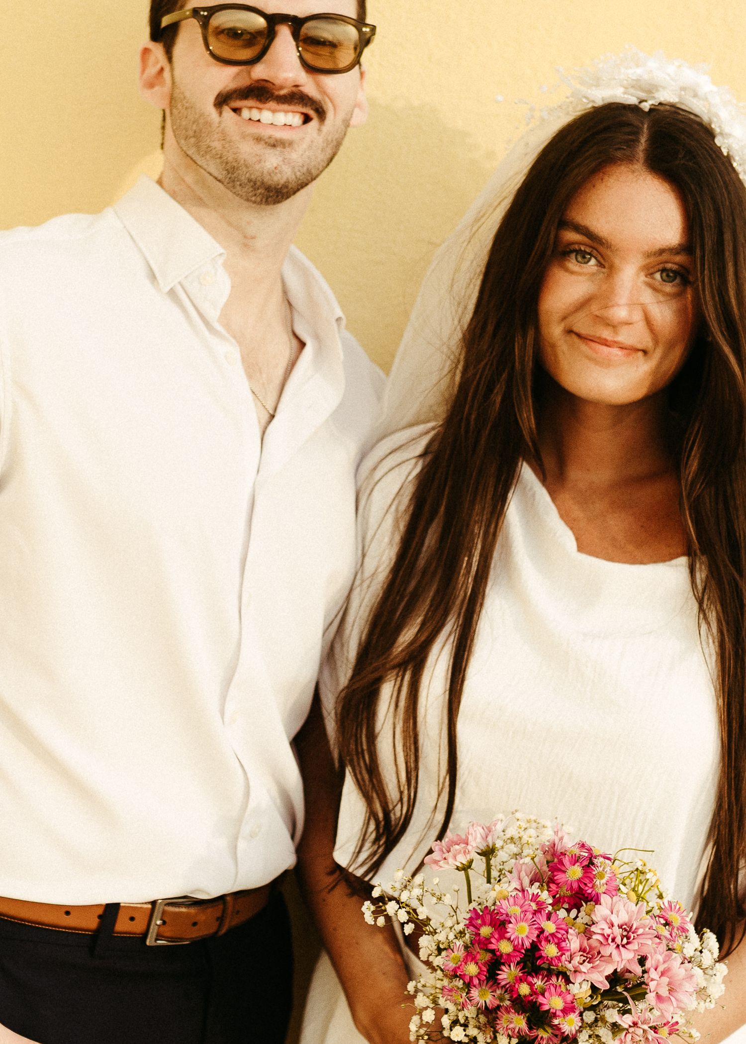 Romantic couple in white attire sharing tender moment with pink floral bouquet.
