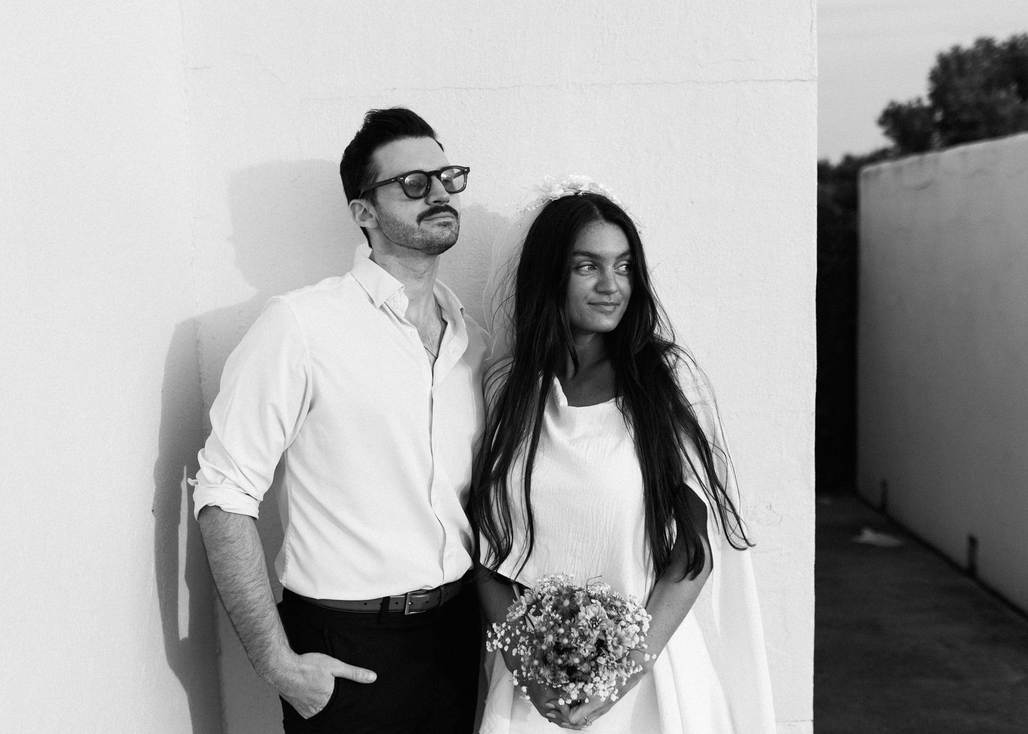 Black and white photo of couple against wall with simple floral arrangement.