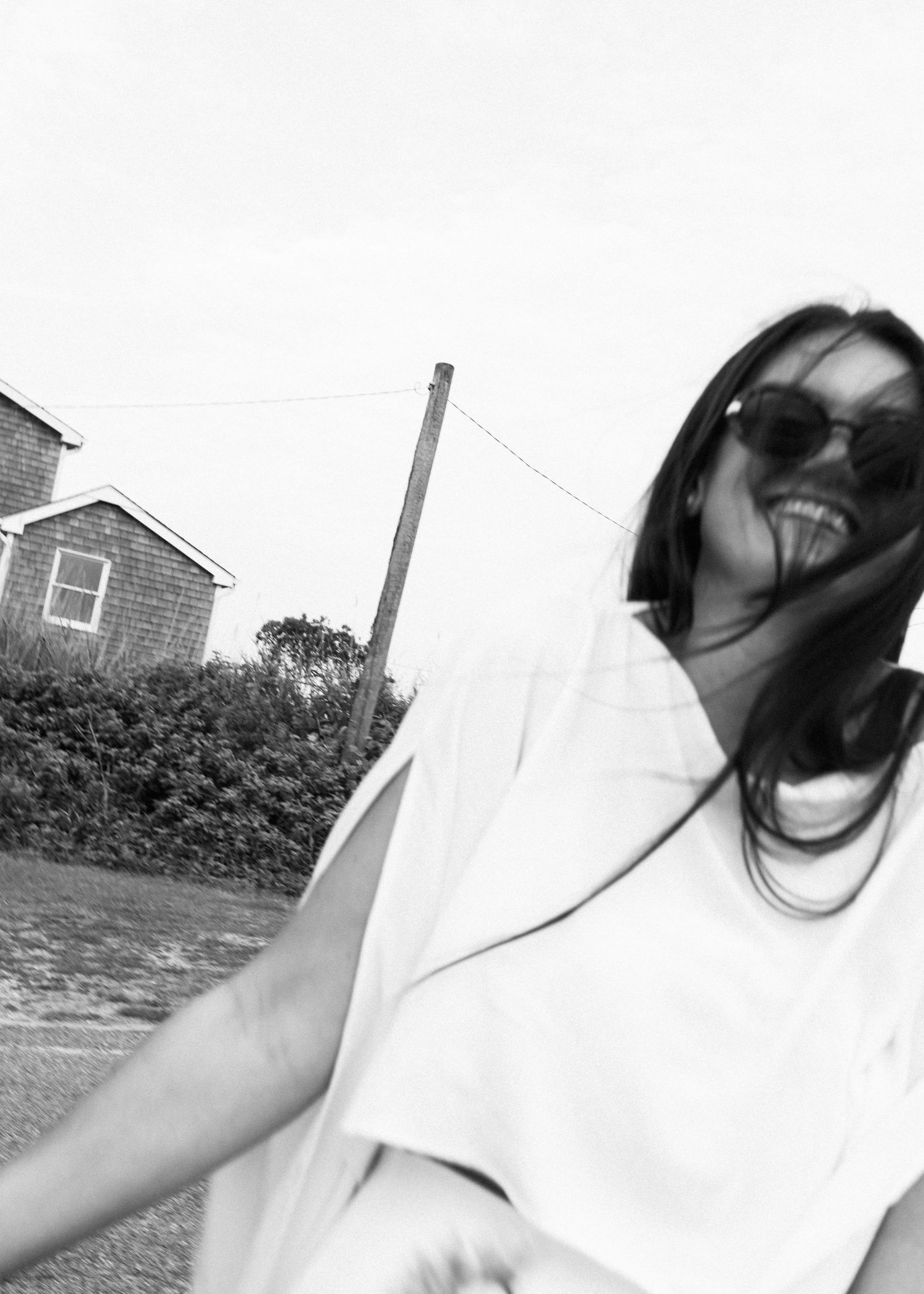A candid black and white shot of someone laughing in sunglasses near a house.