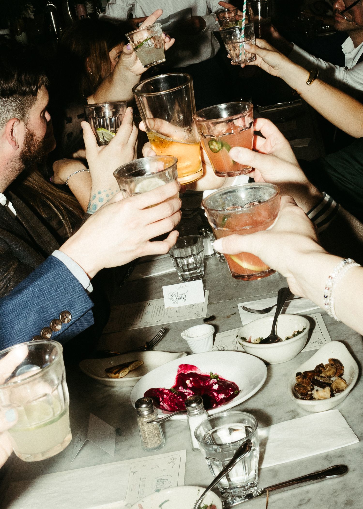 Friends toasting with various cocktails and drinks over a dinner table with food.