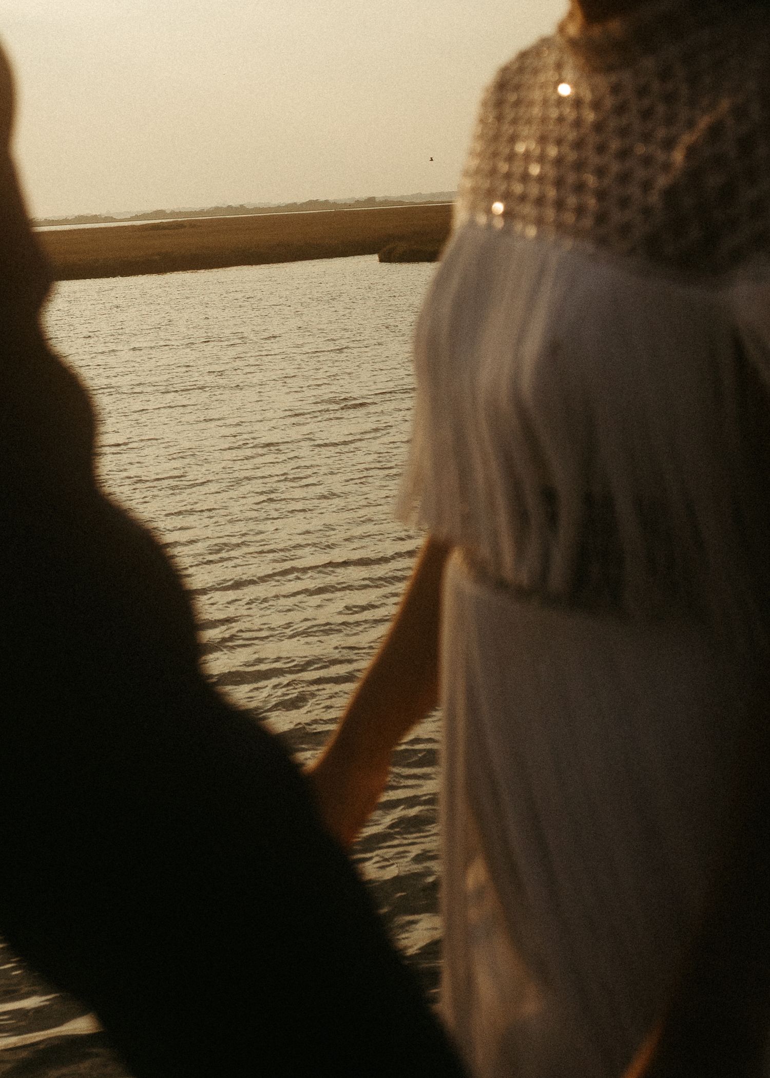 A silhouetted couple holds hands while looking out at a calm ocean during golden hour.