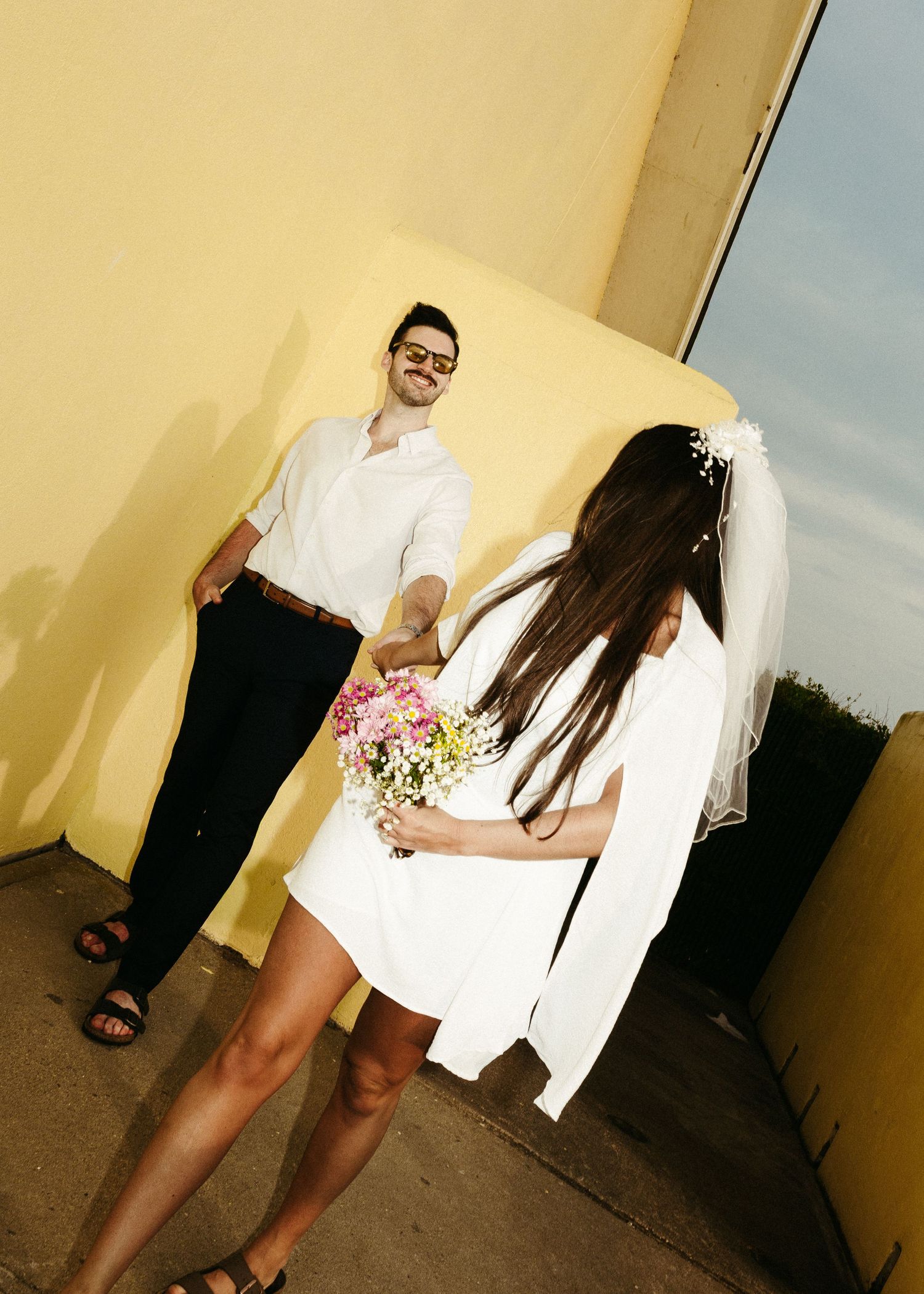 A stylish couple dressed in white poses against a yellow wall during their wedding photoshoot.