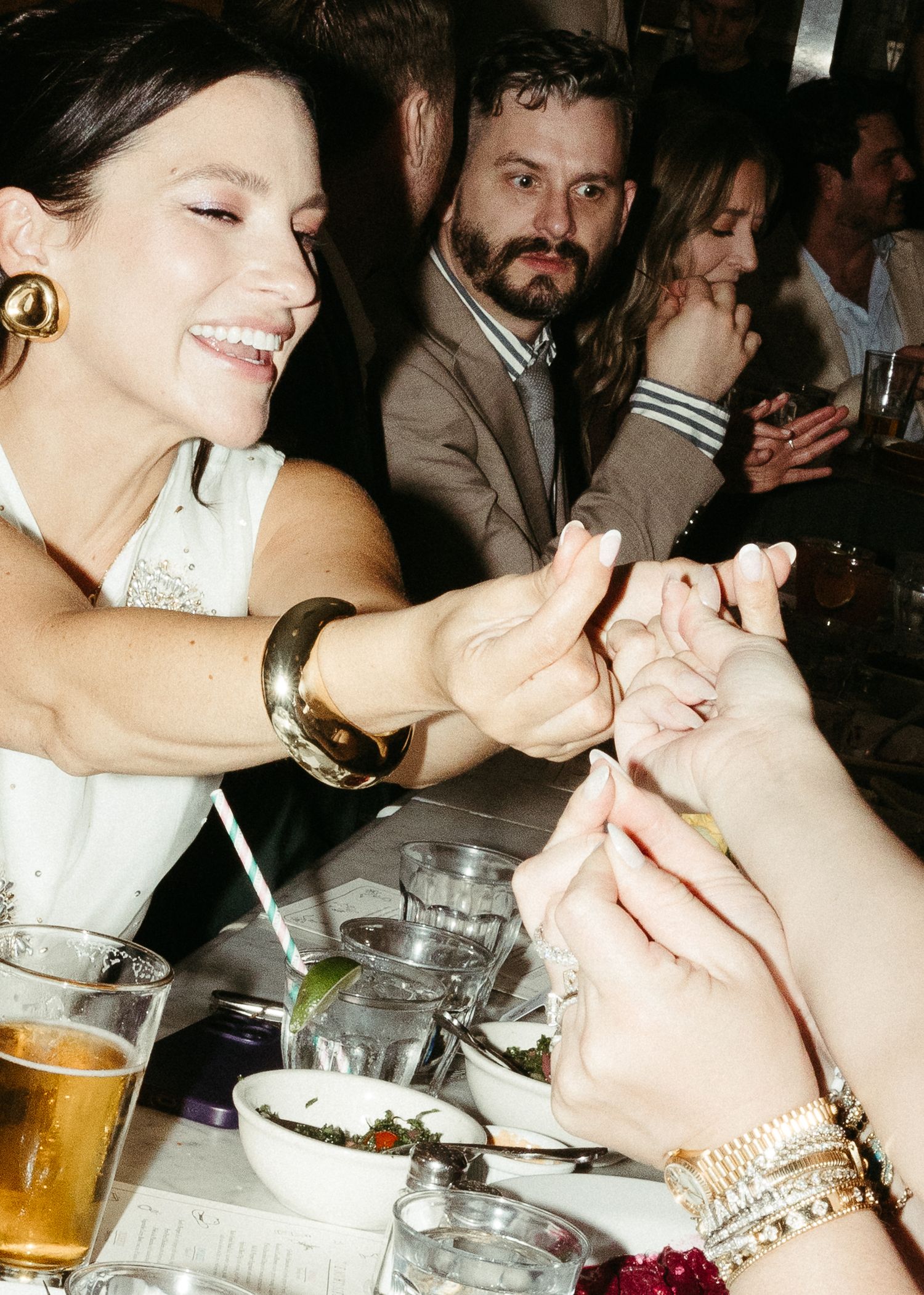 A lively dinner party scene with people laughing and holding hands across a table with drinks and plates.