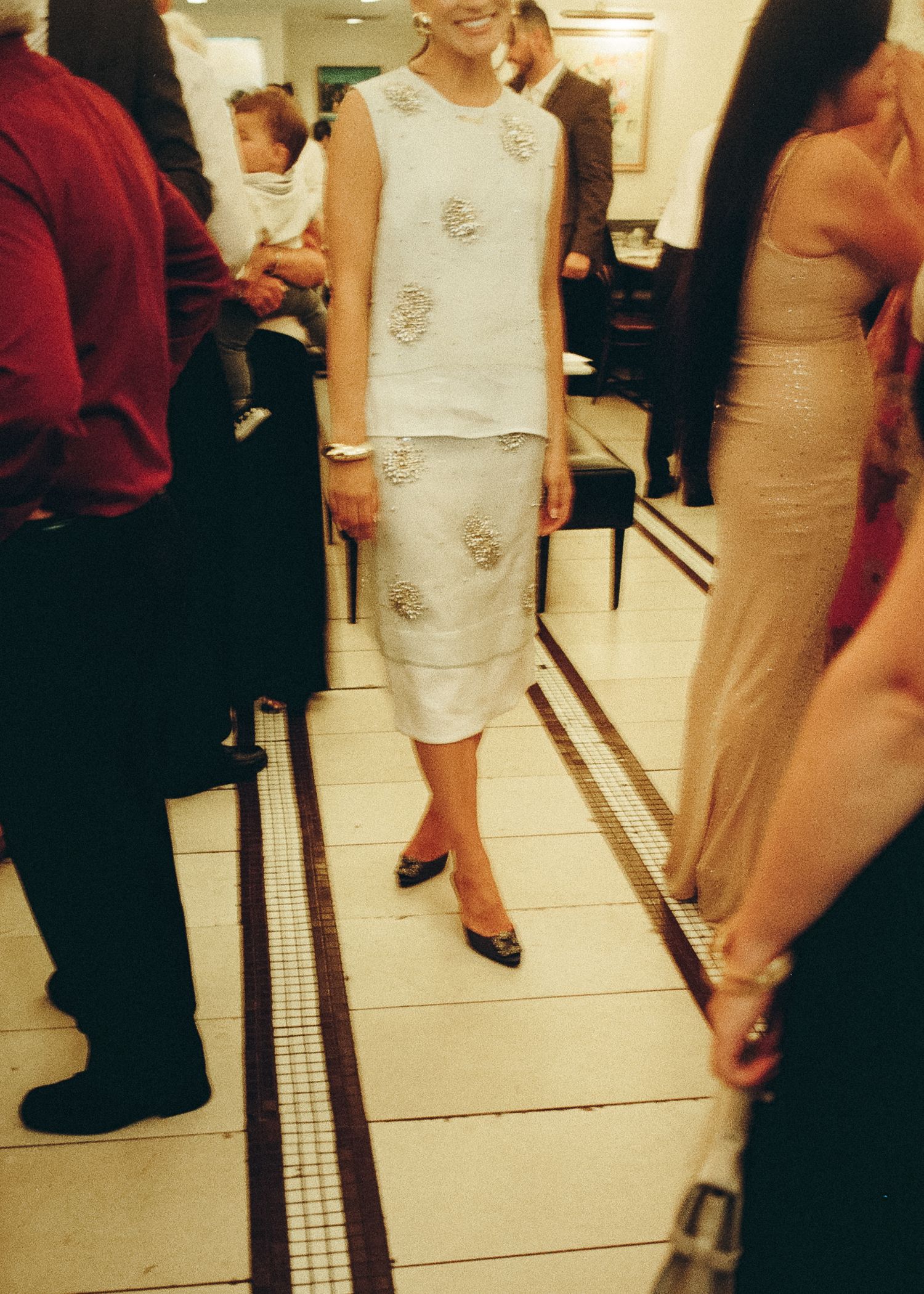Person wearing a white embellished sleeveless dress standing on tiled floor at rehearsal dinner.