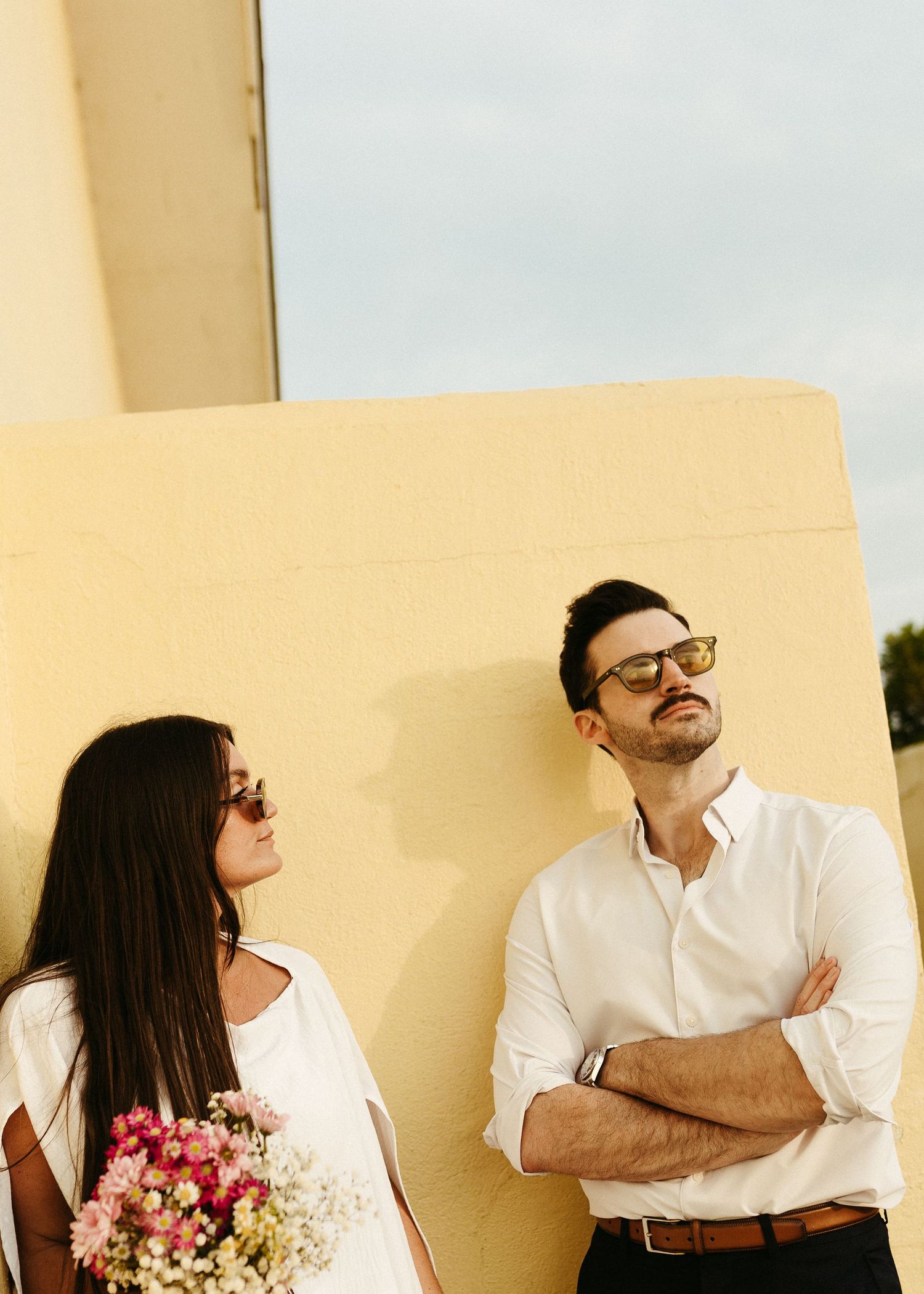 A fashionable couple in white clothing poses against a yellow wall with pink flowers.