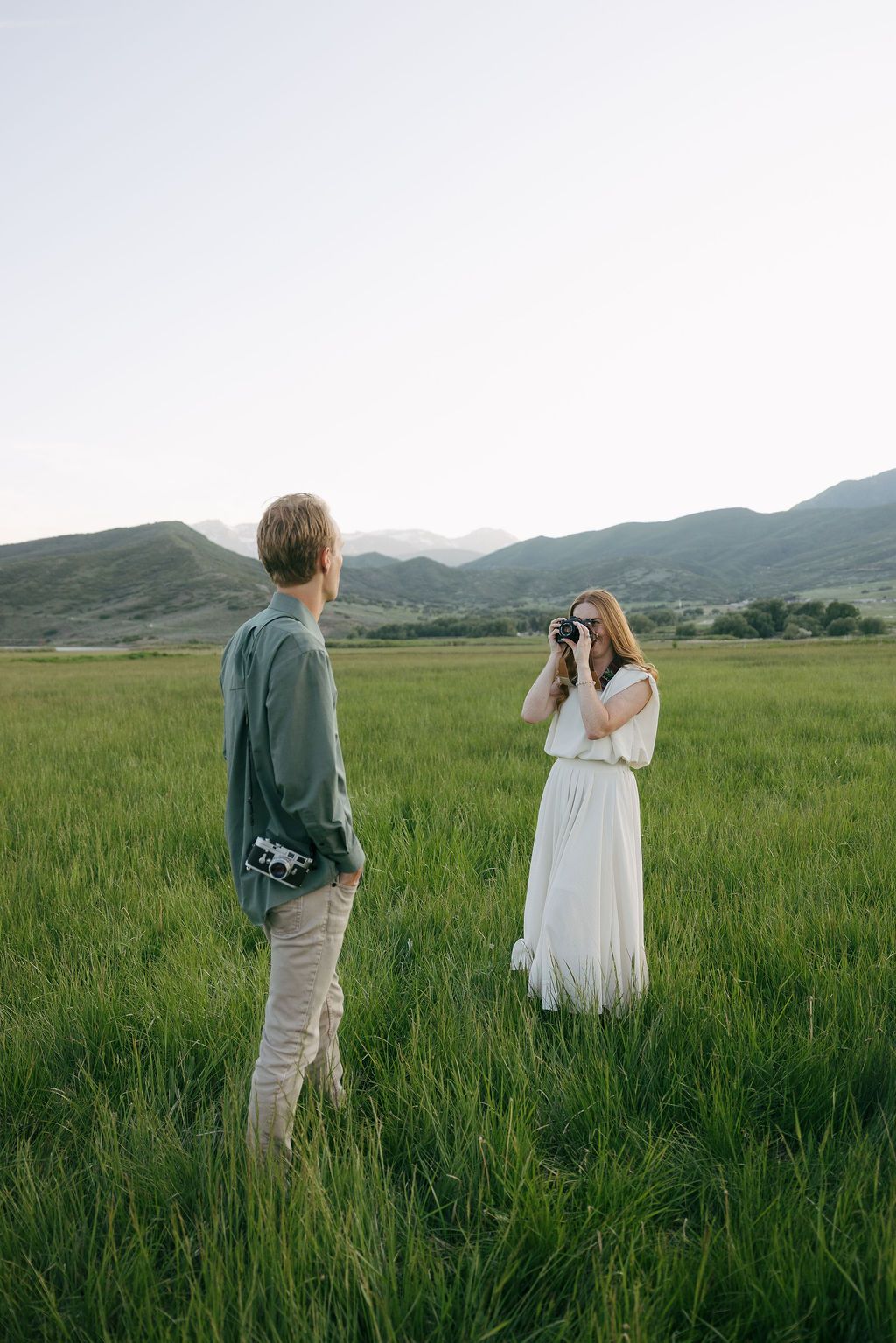 A couple stands together in a lush green meadow with rolling hills in the background at sunset.