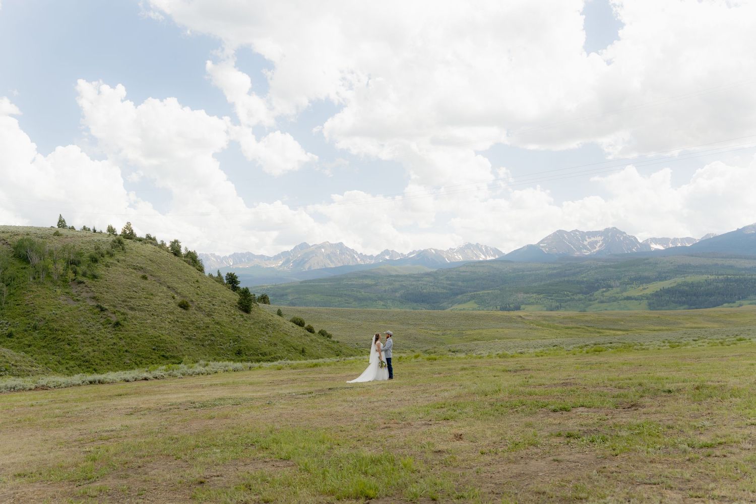 A panoramic view of a couple on their wedding day standing on a grassy hillside with mountains in the distance.