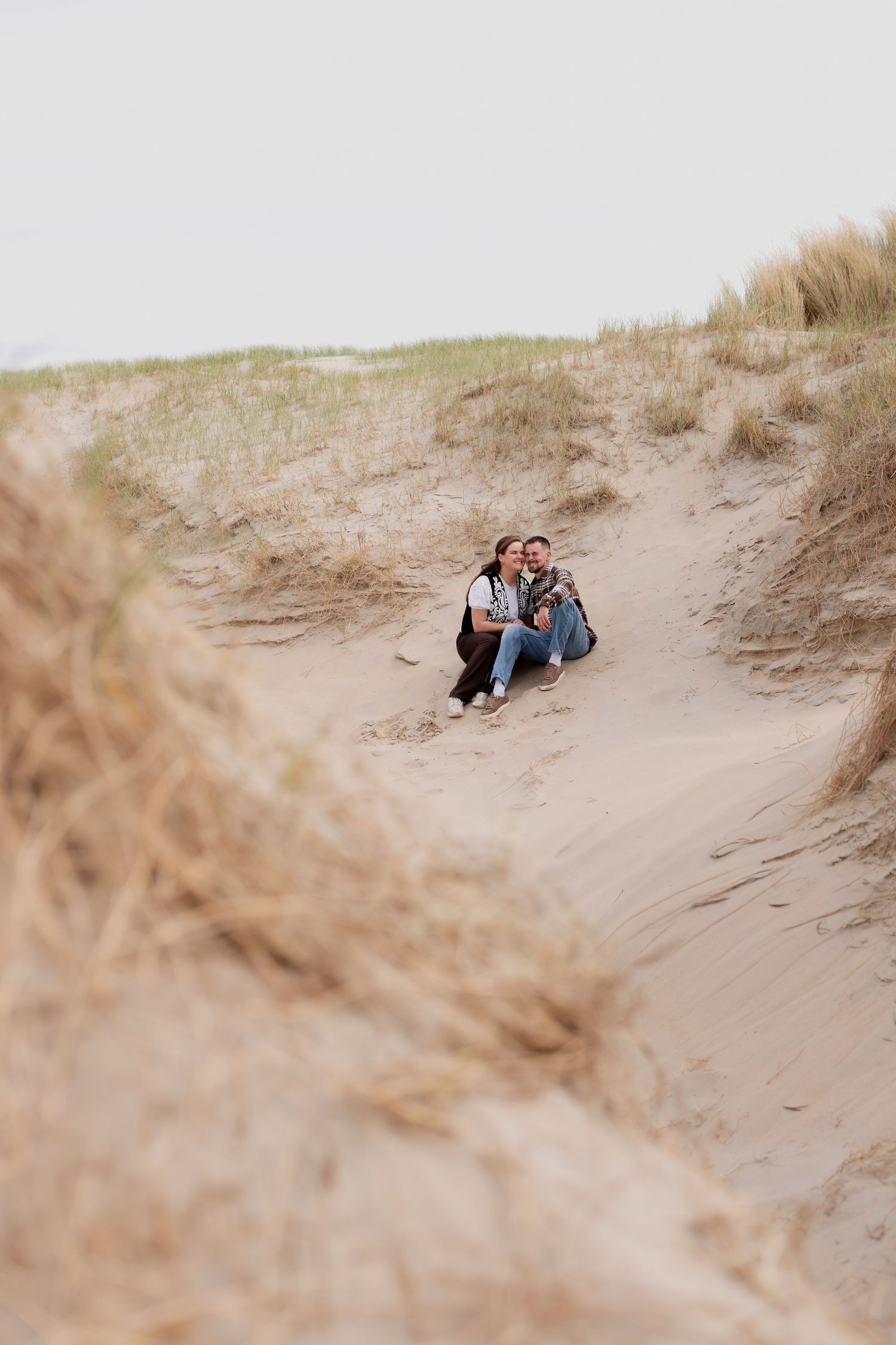 Couple sitting together on sandy beach dunes surrounded by tall grass on an overcast day.