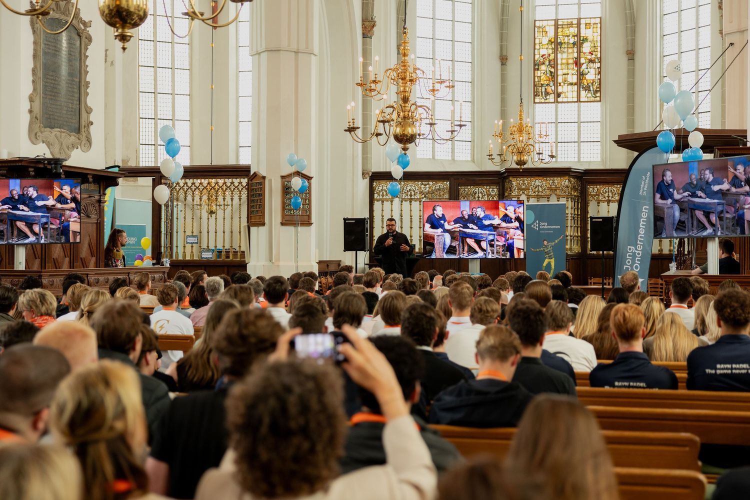 Groot publiek zit in een historische kerk en kijkt naar een presentatie op meerdere schermen tijdens een jongerenevenement.