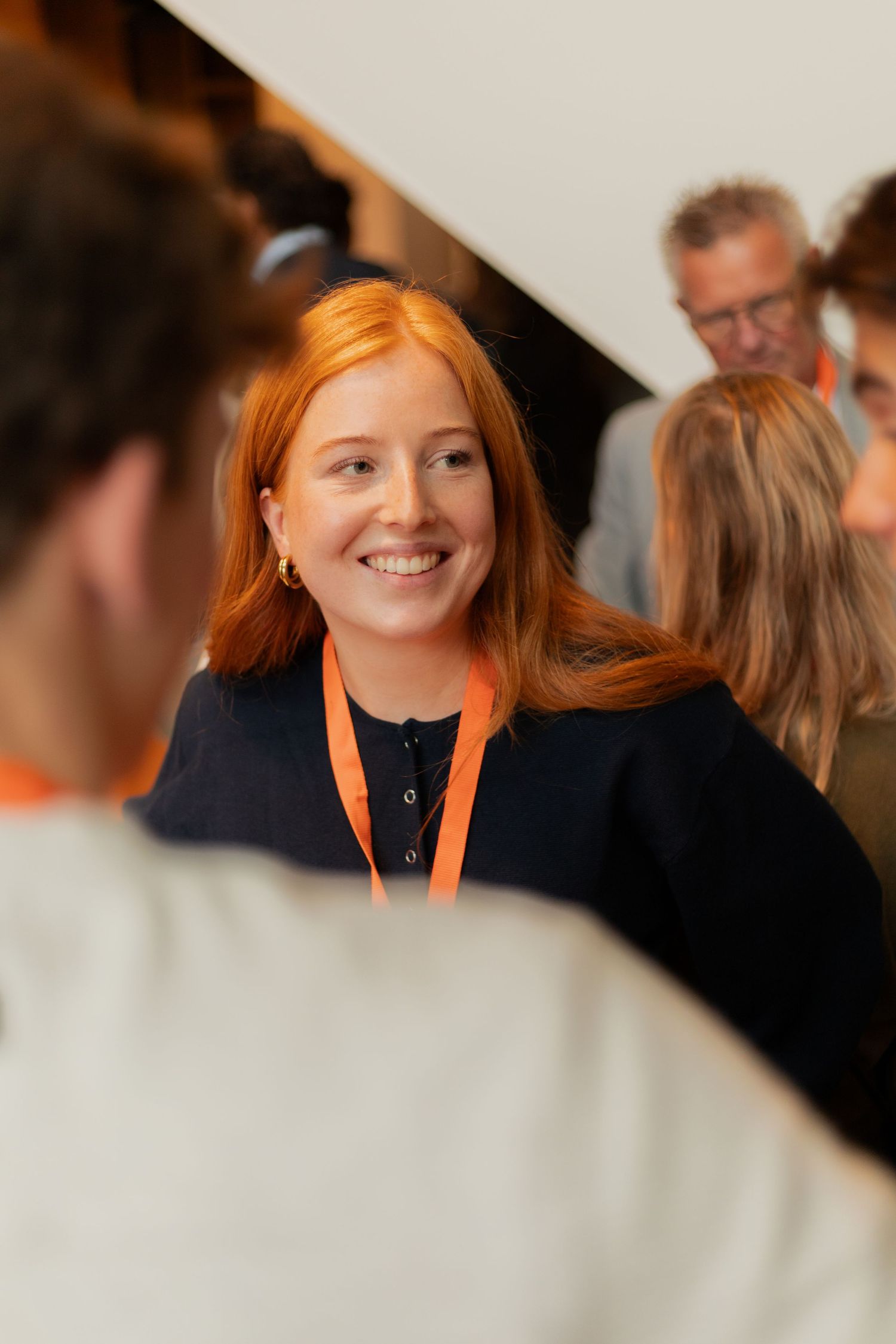 A person with vibrant orange hair wearing a black top and orange lanyard smiles at a professional networking event.