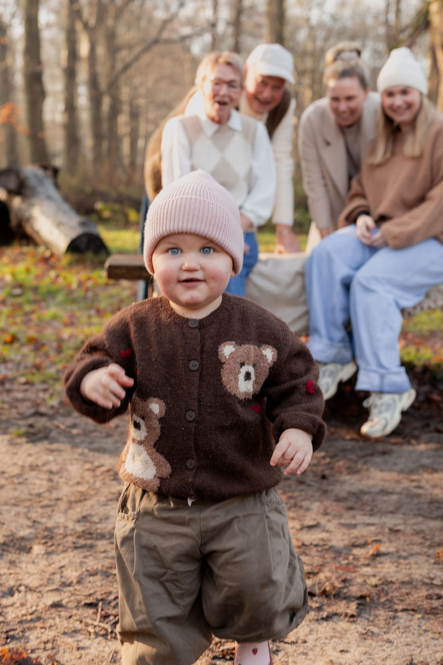 A small child wearing a brown bear sweater and pink beanie stands on a forest path during autumn while others sit behind.